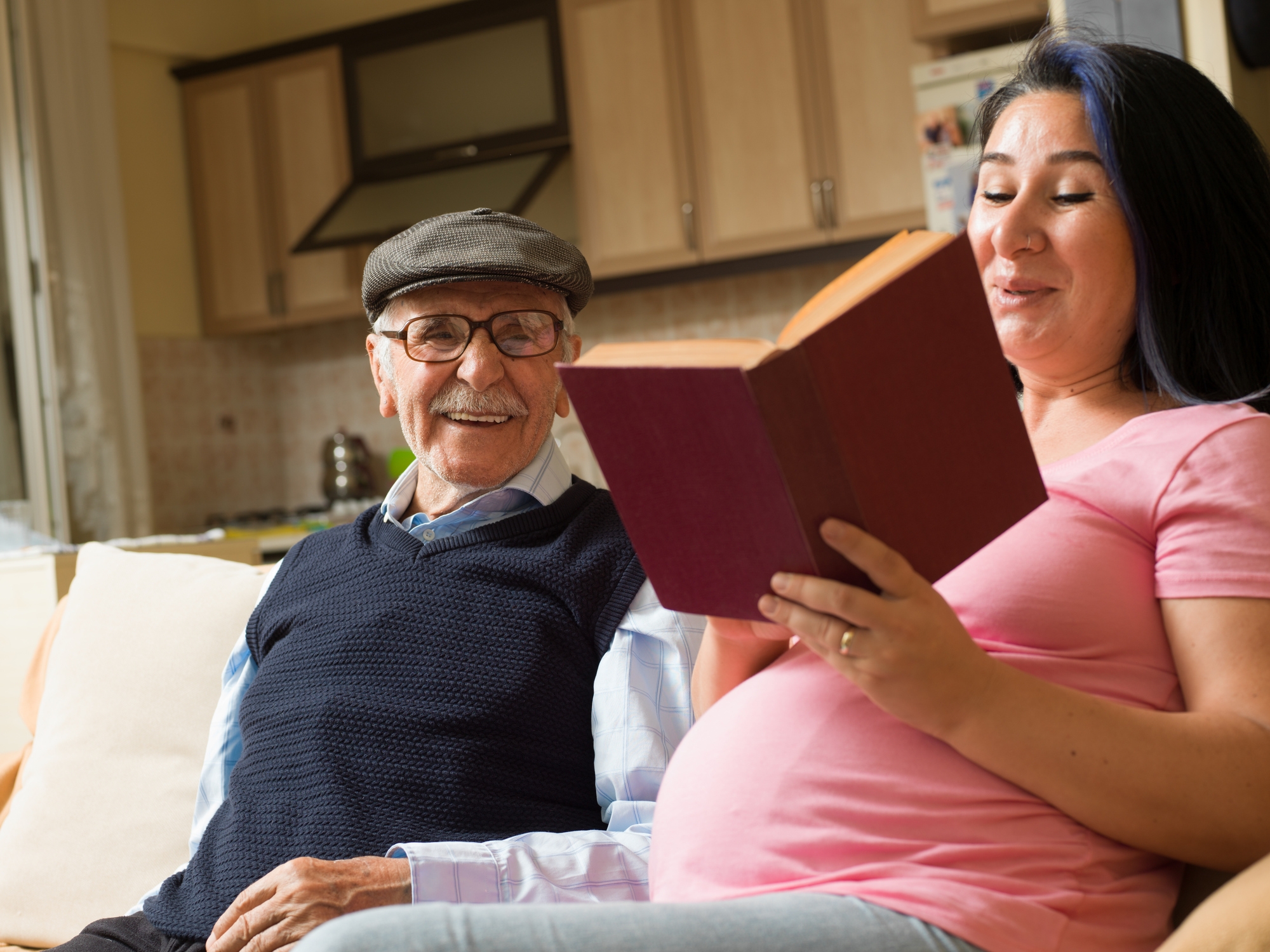 An elderly man and a pregnant woman sit on a couch; she reads a book aloud while he smiles, both appearing relaxed and content in a home setting