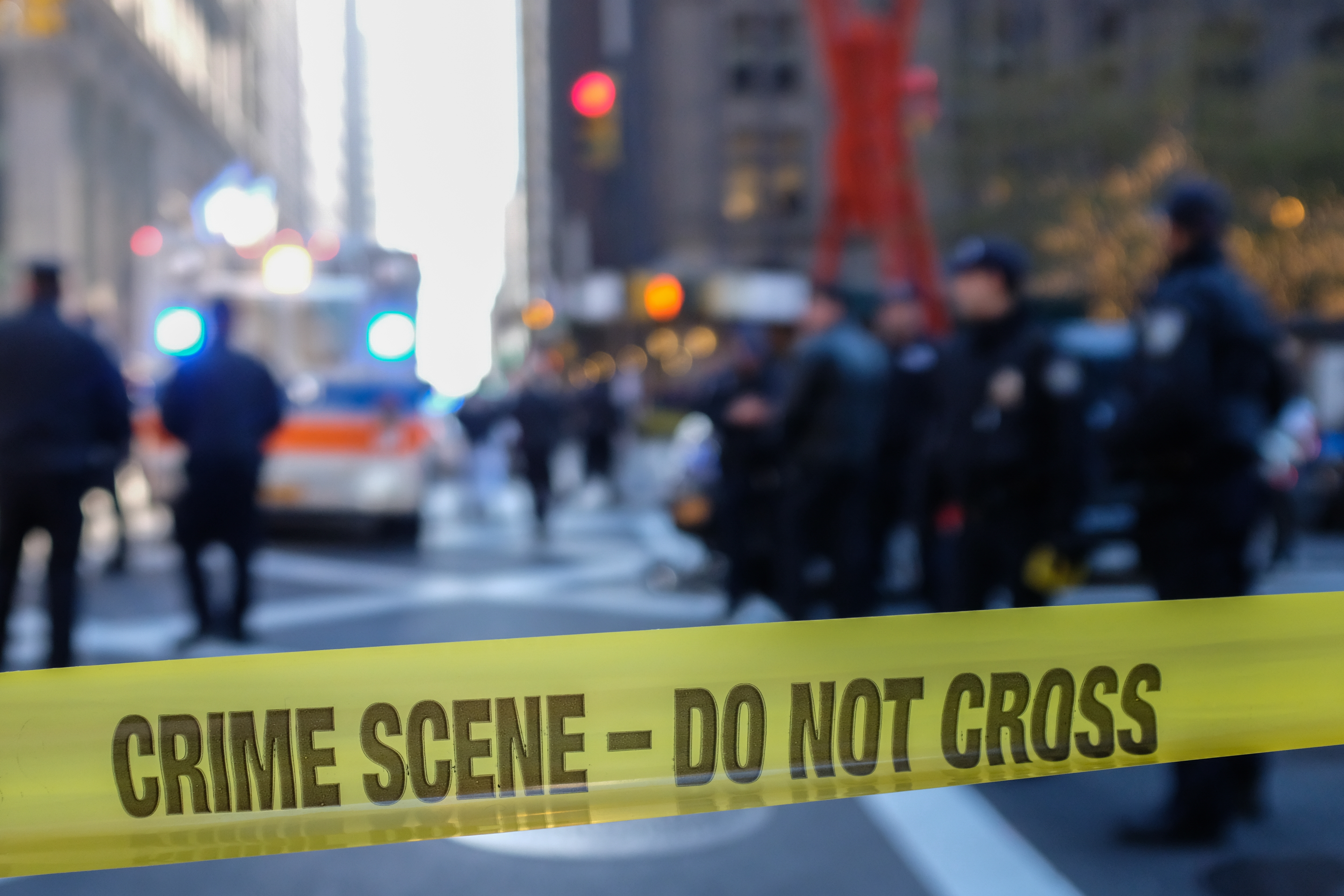 Blurred city street scene with focused yellow crime scene tape reading "CRIME SCENE - DO NOT CROSS" in the foreground
