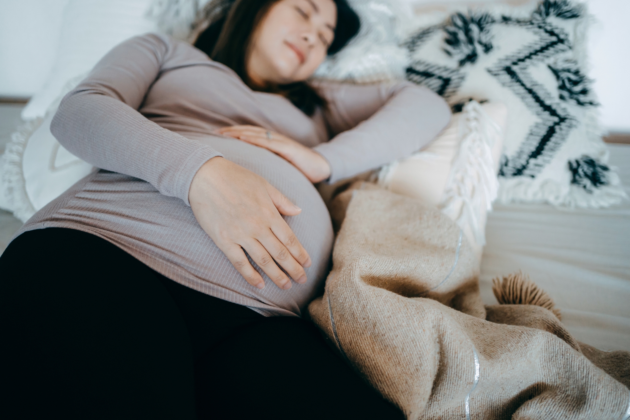 Pregnant person resting on bed with a hand on their stomach, surrounded by cozy pillows and a blanket