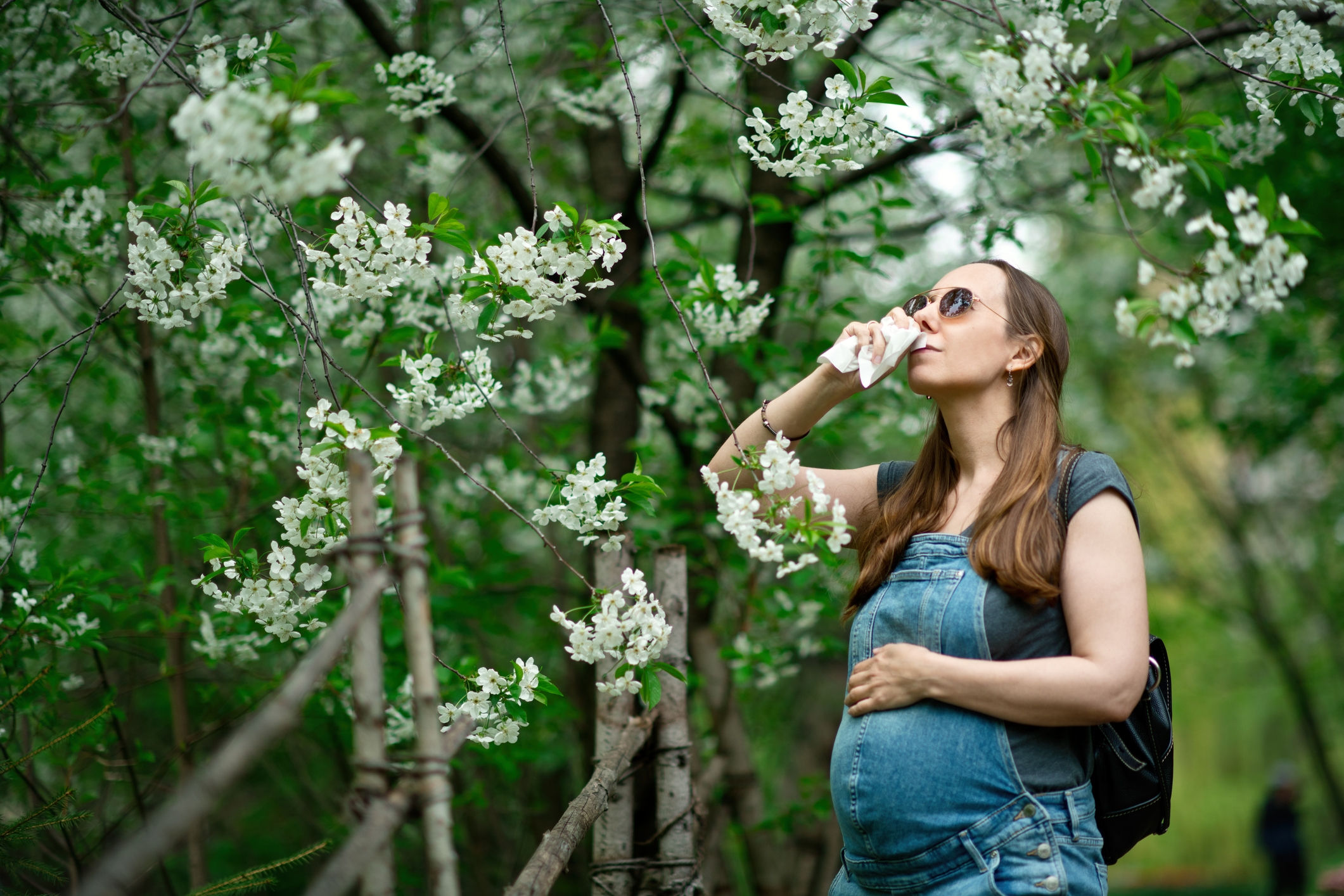 Pregnant woman in overalls uses a tissue while standing under blooming trees, wearing sunglasses and a backpack, enjoying nature