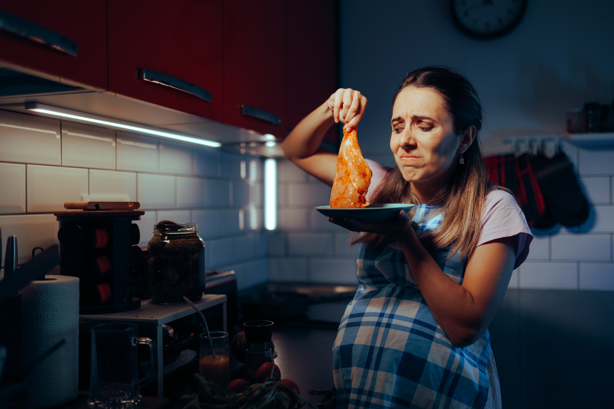 Pregnant woman in kitchen grimacing while holding raw chicken leg on plate, suggesting distaste or discomfort