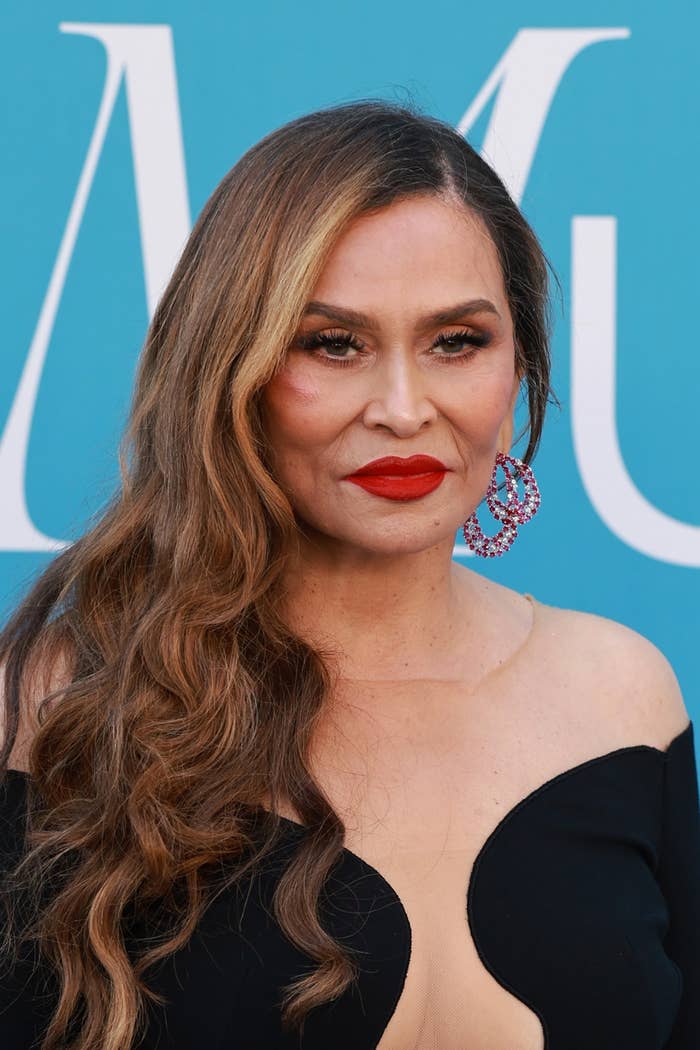 Tina Knowles on a blue background, wearing an off-the-shoulder black dress and large hoop earrings, with long, wavy hair