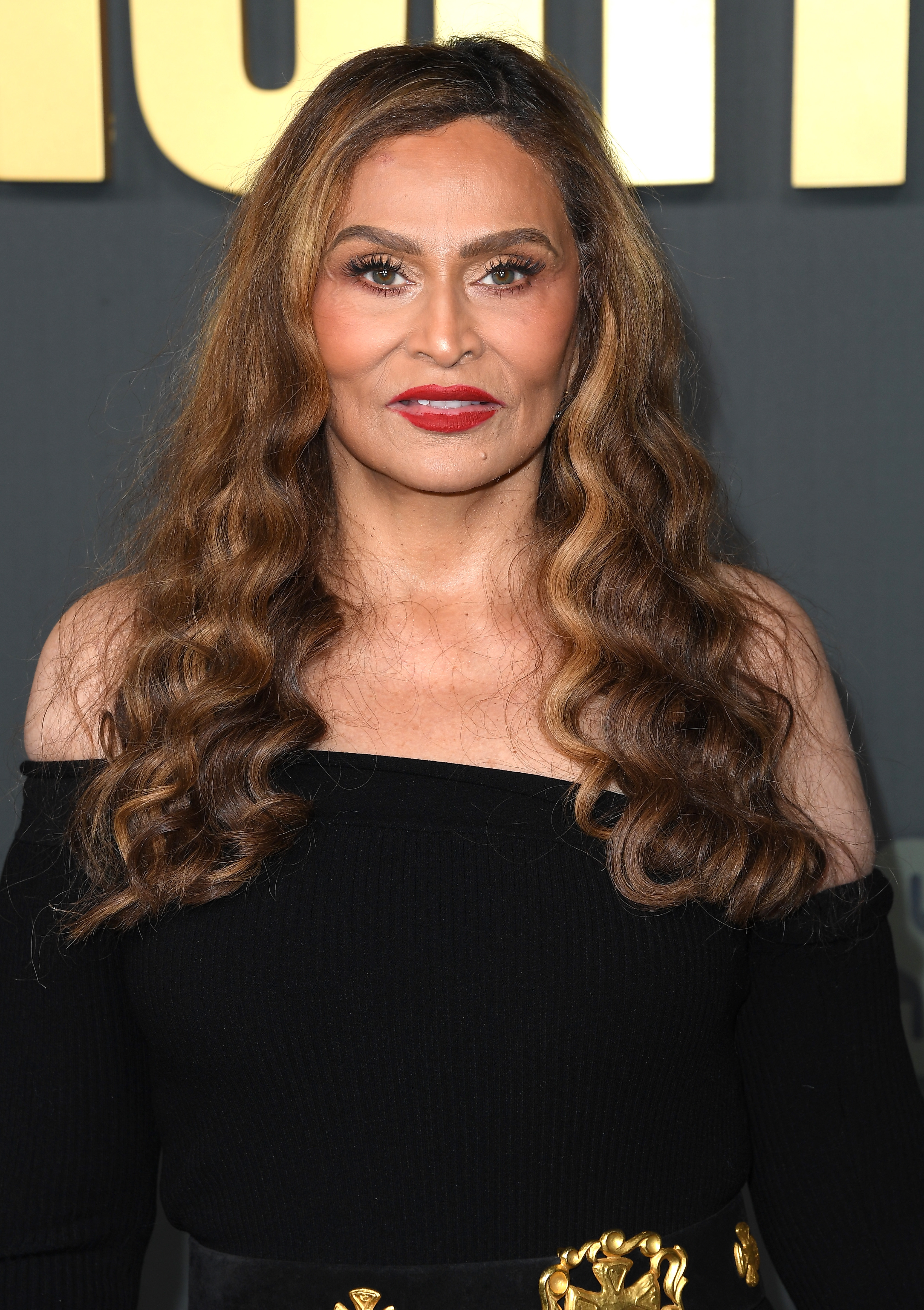 Tina Knowles with long wavy hair, wearing an off-shoulder top and a belt with gold accents, standing in front of a backdrop