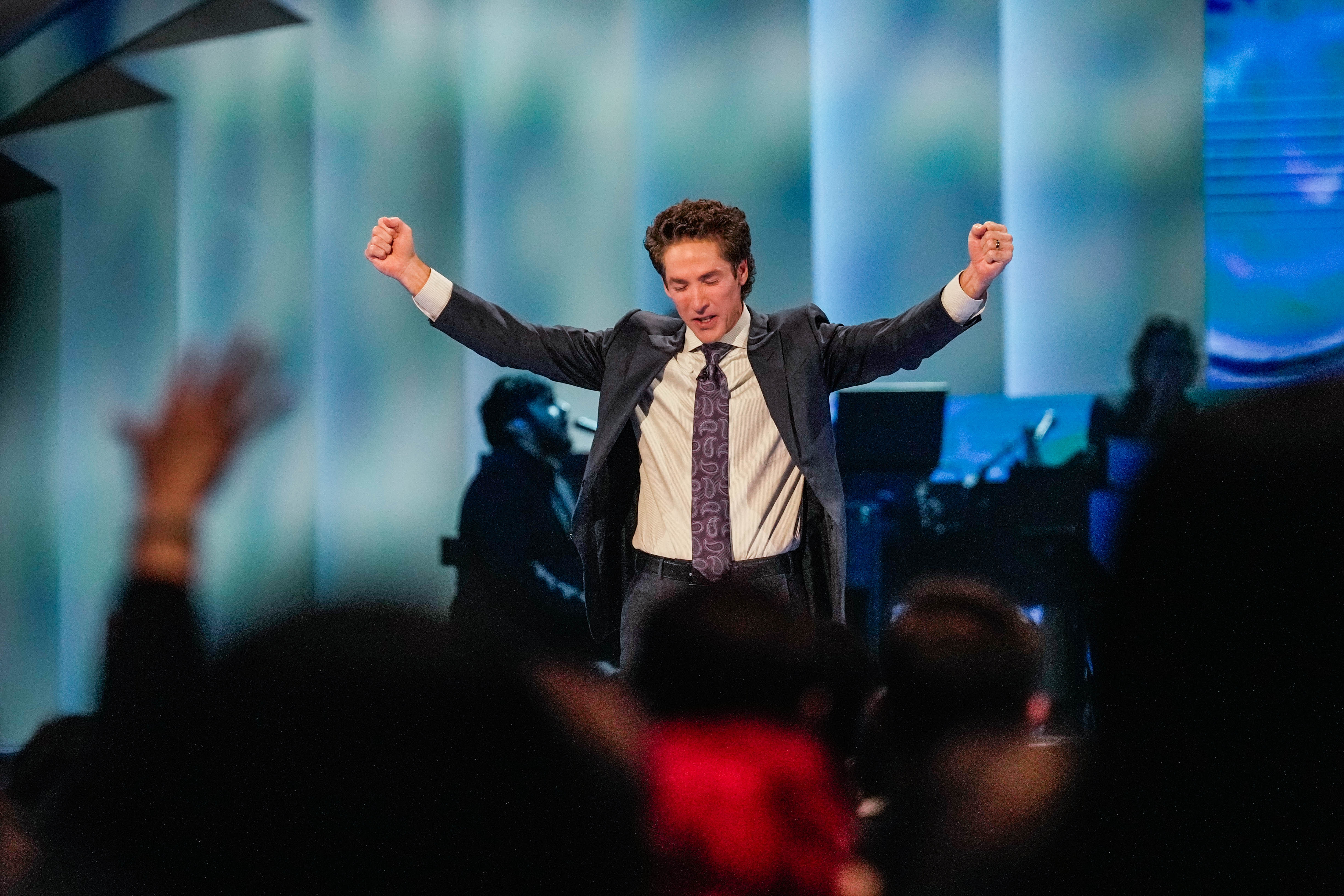 Joel Osteen in a suit with raised arms on stage, speaking to an audience, with a musical group in the background