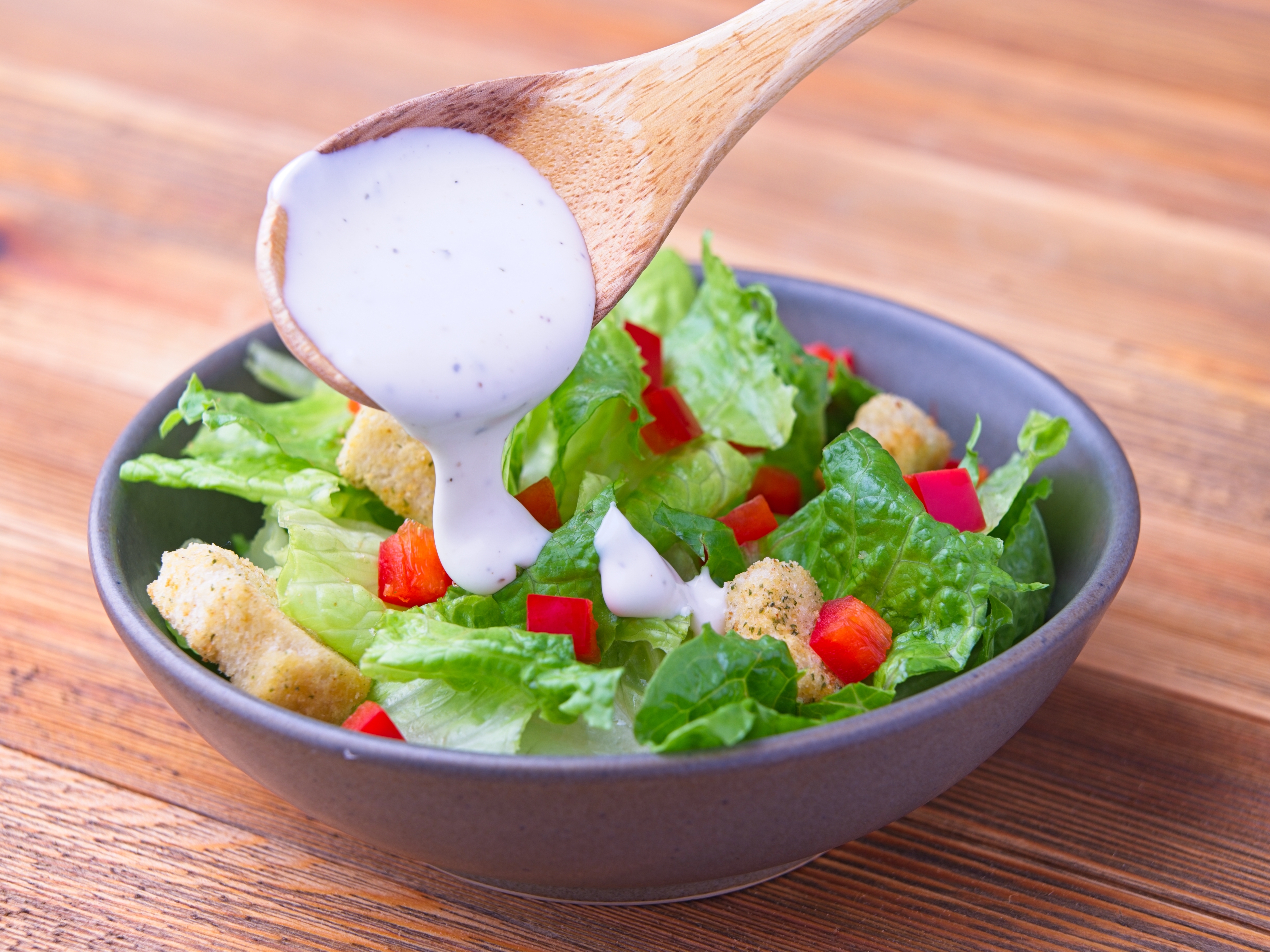 Ranch dressing is being poured over a salad with lettuce, croutons, and diced red peppers in a bowl