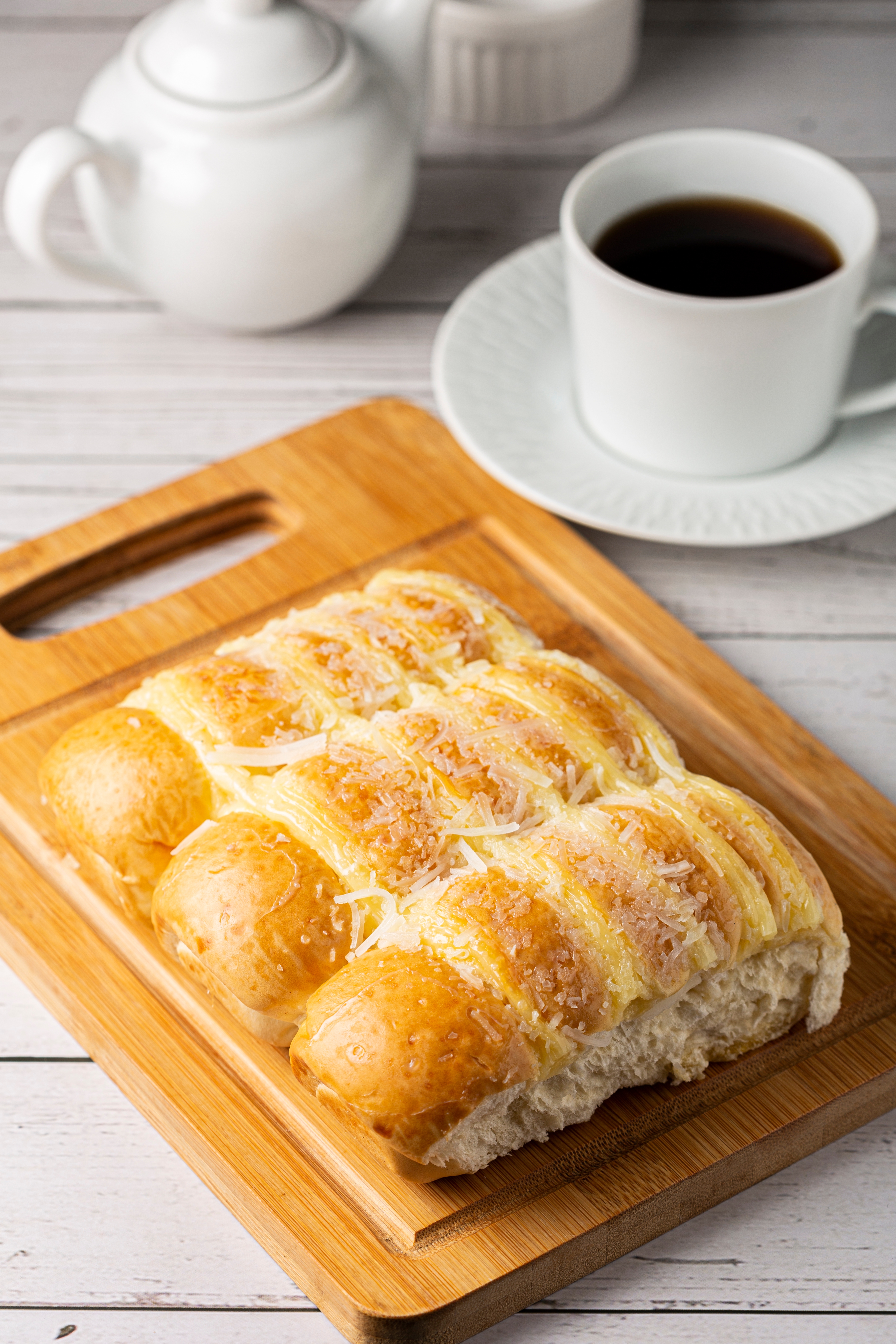 Bread topped with cheese on a wooden board, with a teapot and cup of coffee beside it on a table