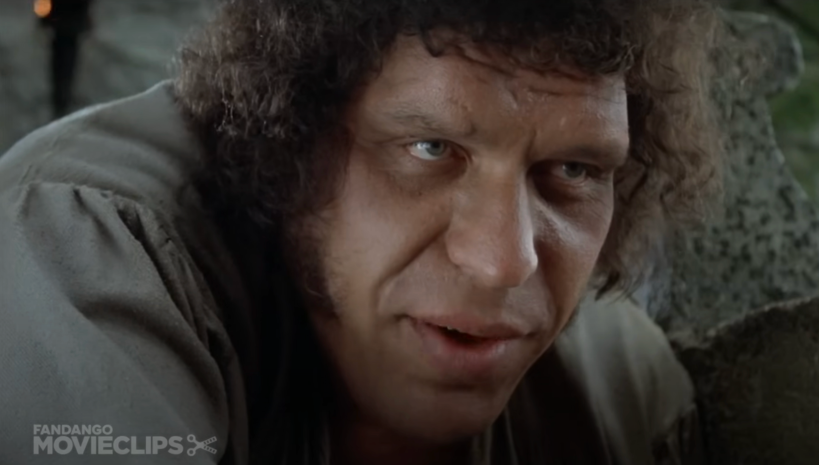 Fezzik with curly hair leans forward, speaking intently. The background is blurred stone