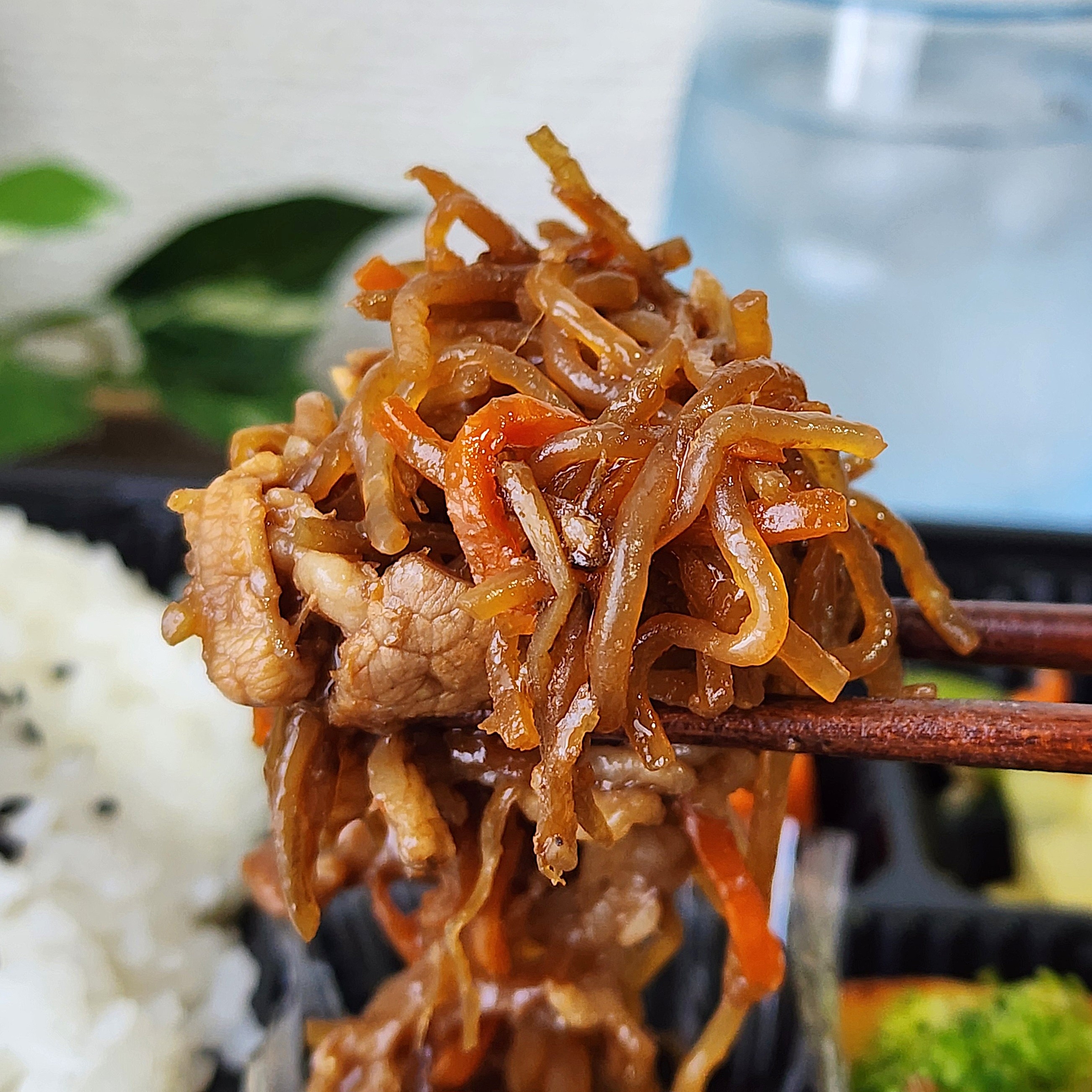 箸で掴まれた肉と野菜の炒め物。後ろに白ごはんと飲み物が見える食事シーン。