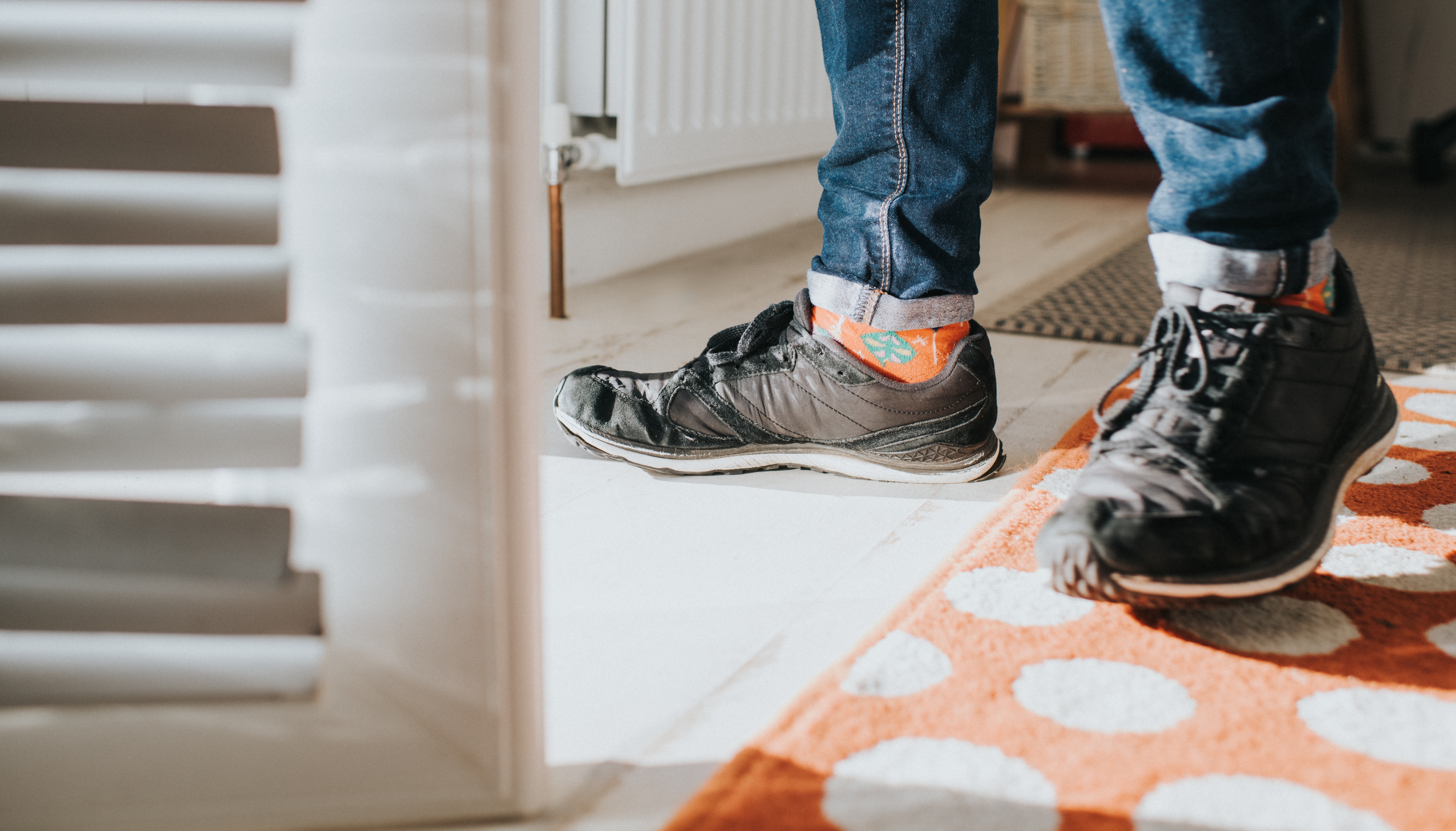 Person wearing worn sneakers and colorful socks stands on a polka dot rug near a window with blinds