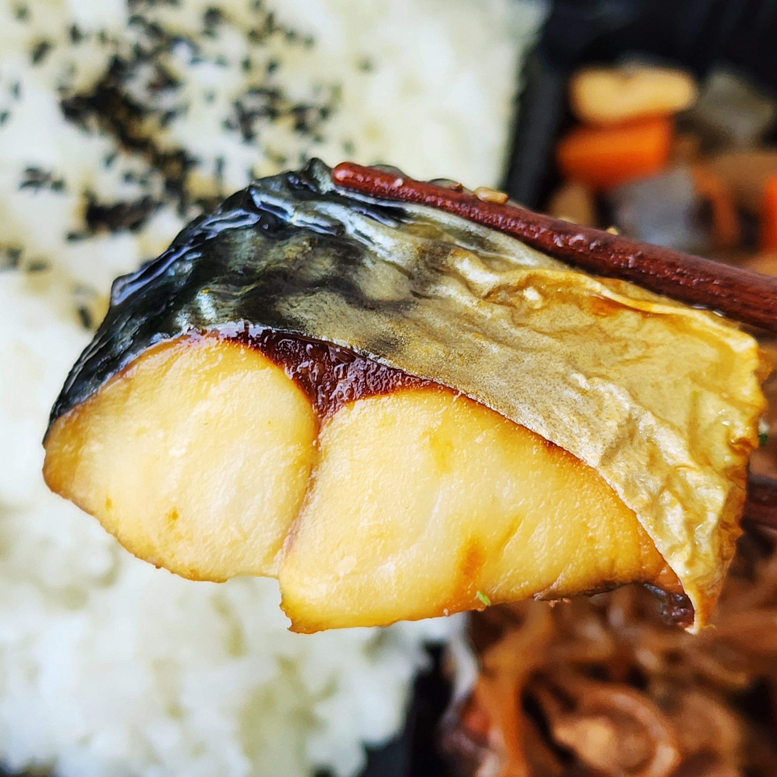 箸で持ち上げられた焼き魚の一切れ。背景に白いご飯と野菜の煮物があります。