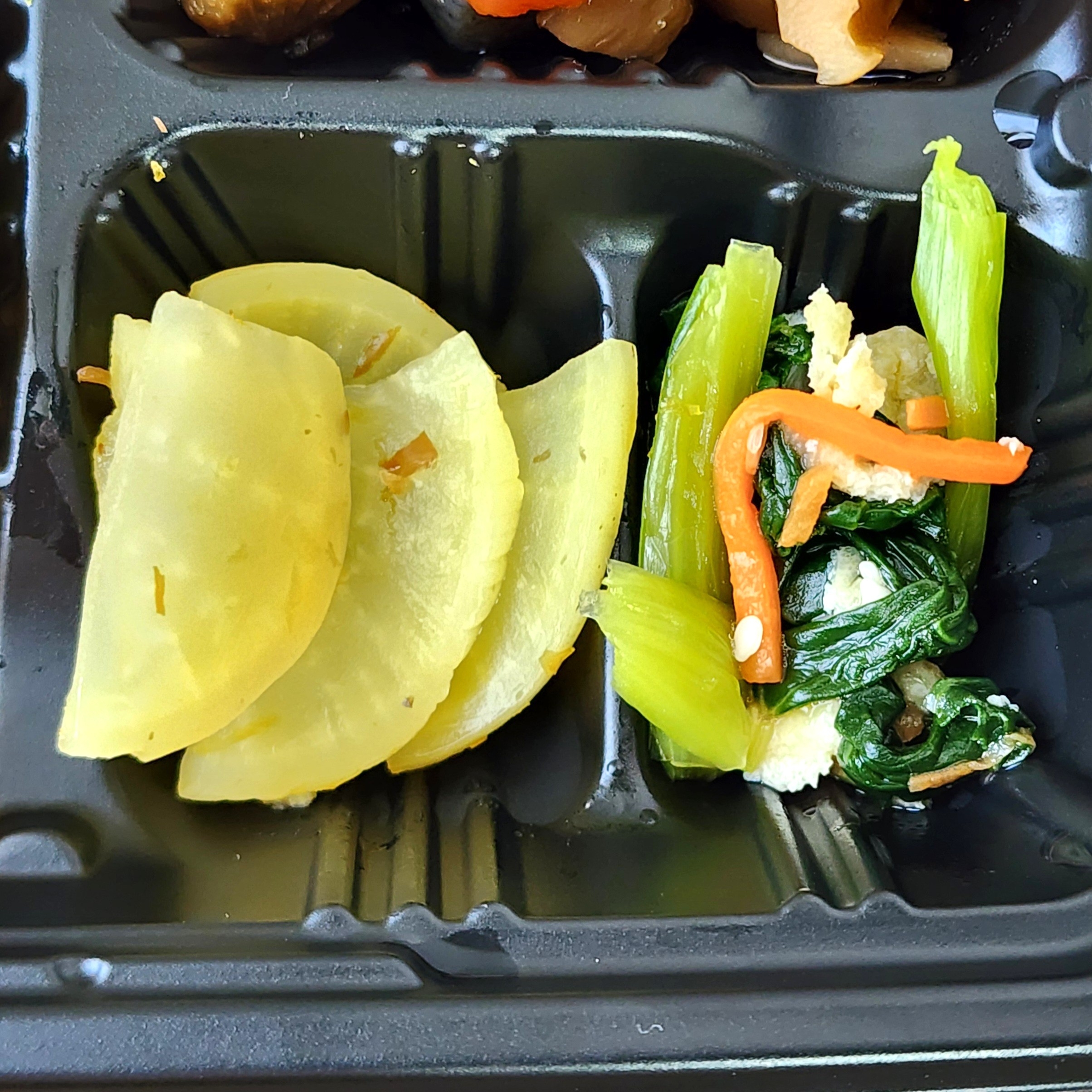 弁当の一部で、大根の漬物とほうれん草、ニンジン、ネギの炒め物がプラスチック容器に入っている。