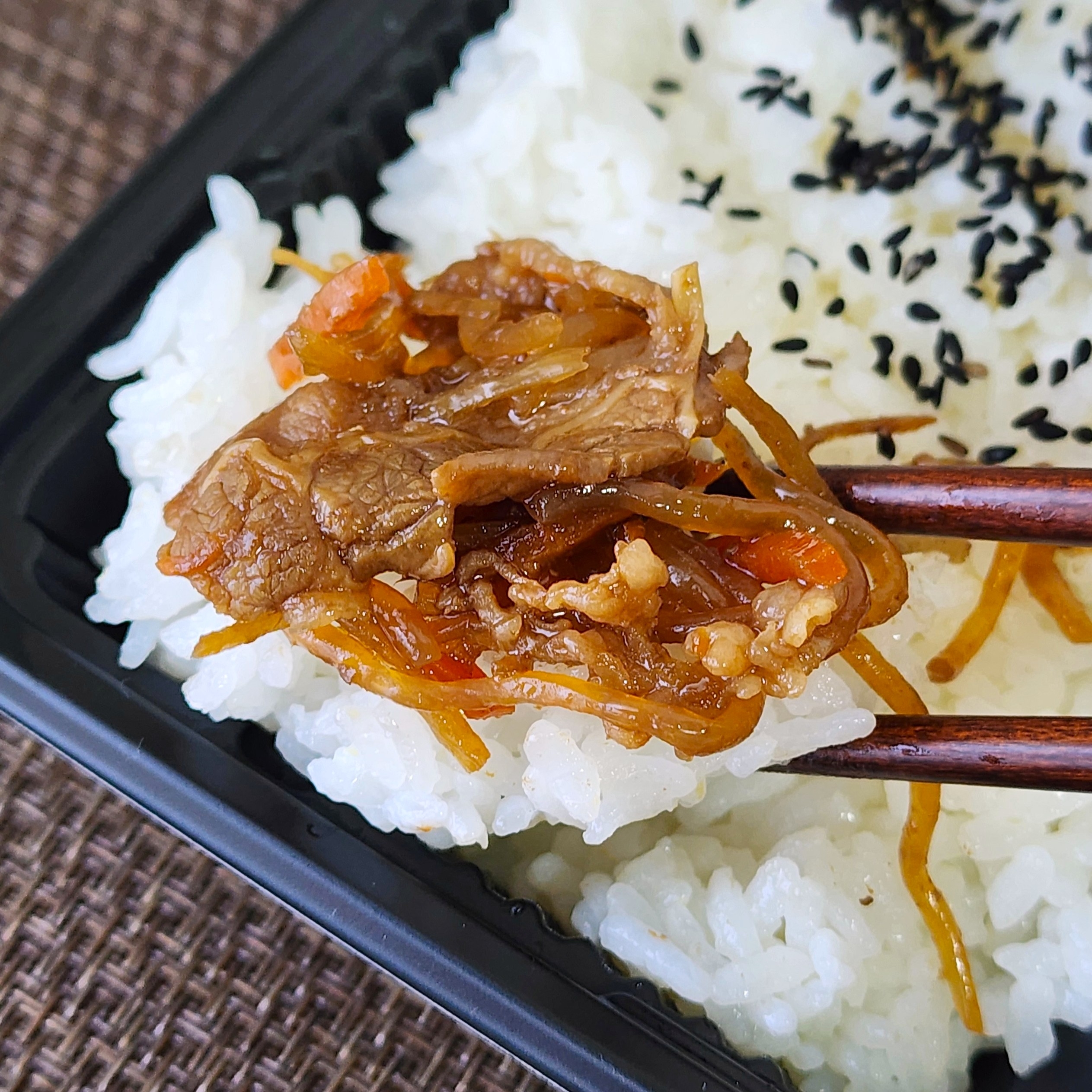 箸で白いご飯と肉の炒め物が食べられている場面。ゴマがご飯の上にトッピングされています。