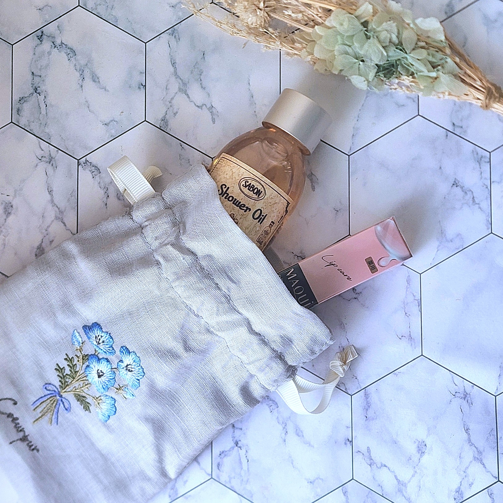 大理石模様の背景に、刺繍入りの布袋に入ったシャワーオイルとリップグロスが見える。隣にドライフラワーの飾りがある。