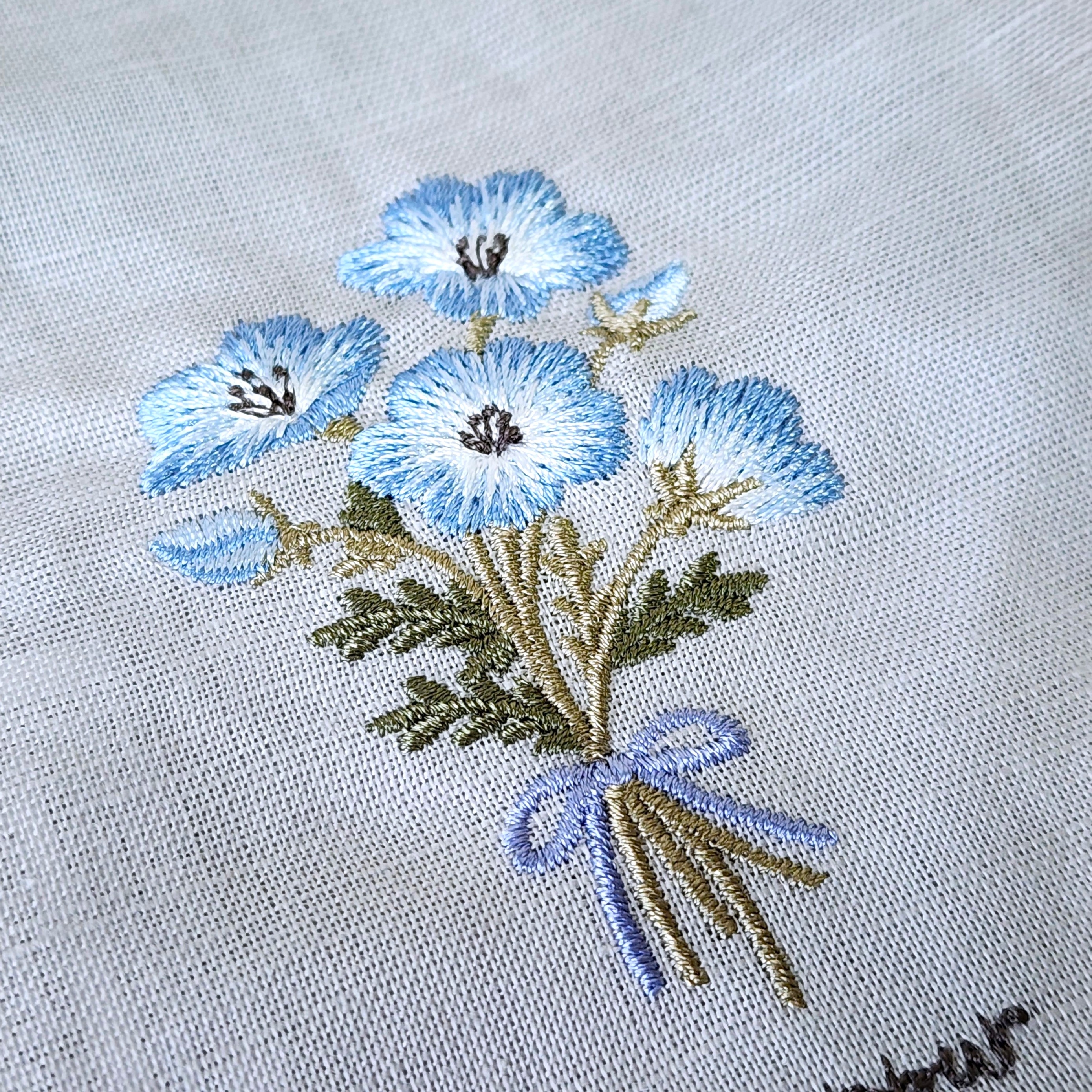 布地に刺繍された花束デザイン。青い花と葉がリアルに表現され、リボンで結ばれた繊細な作品。