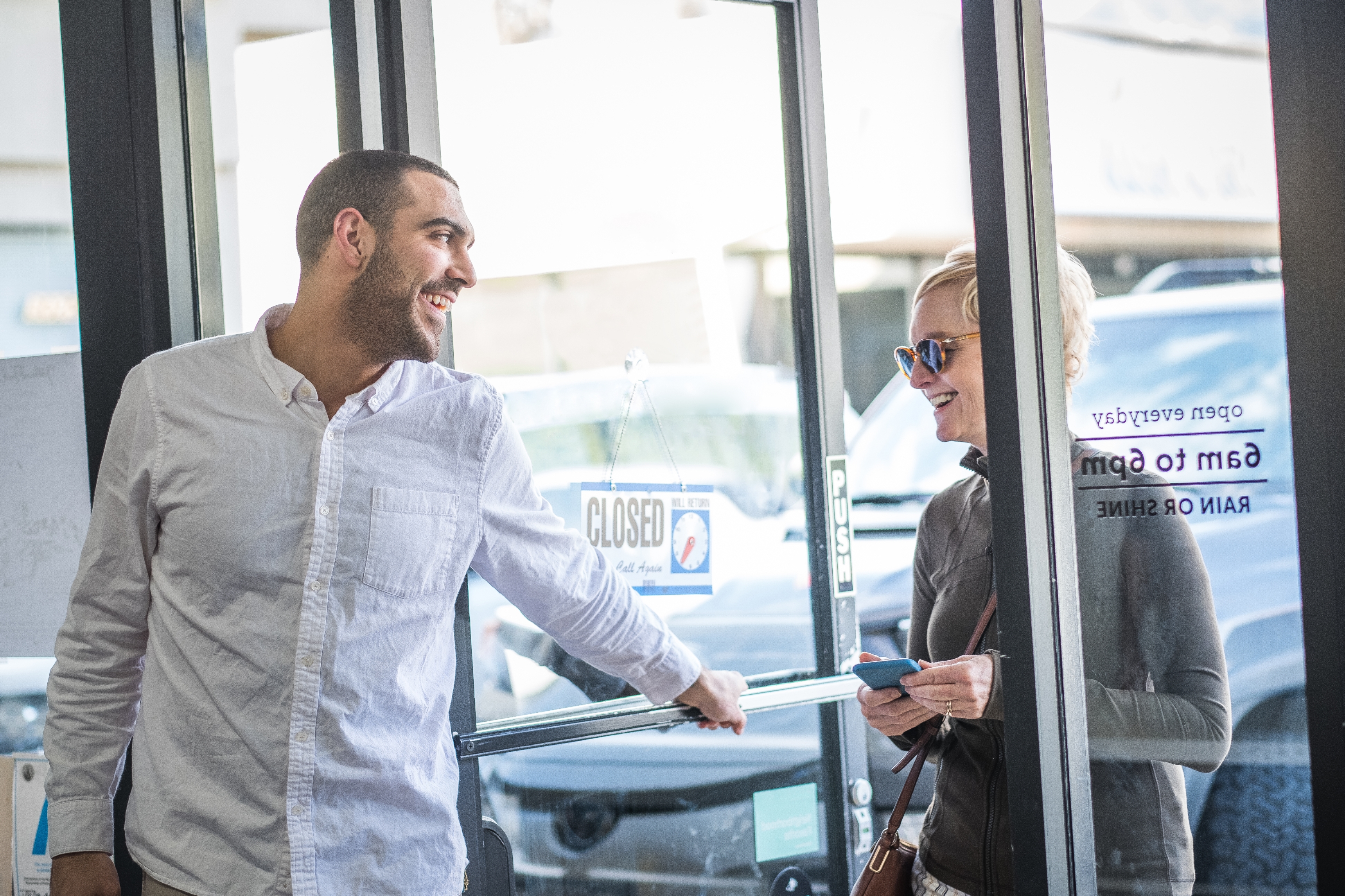 Man and woman smiling at each other at a shop entrance; the door displays an open/closed sign