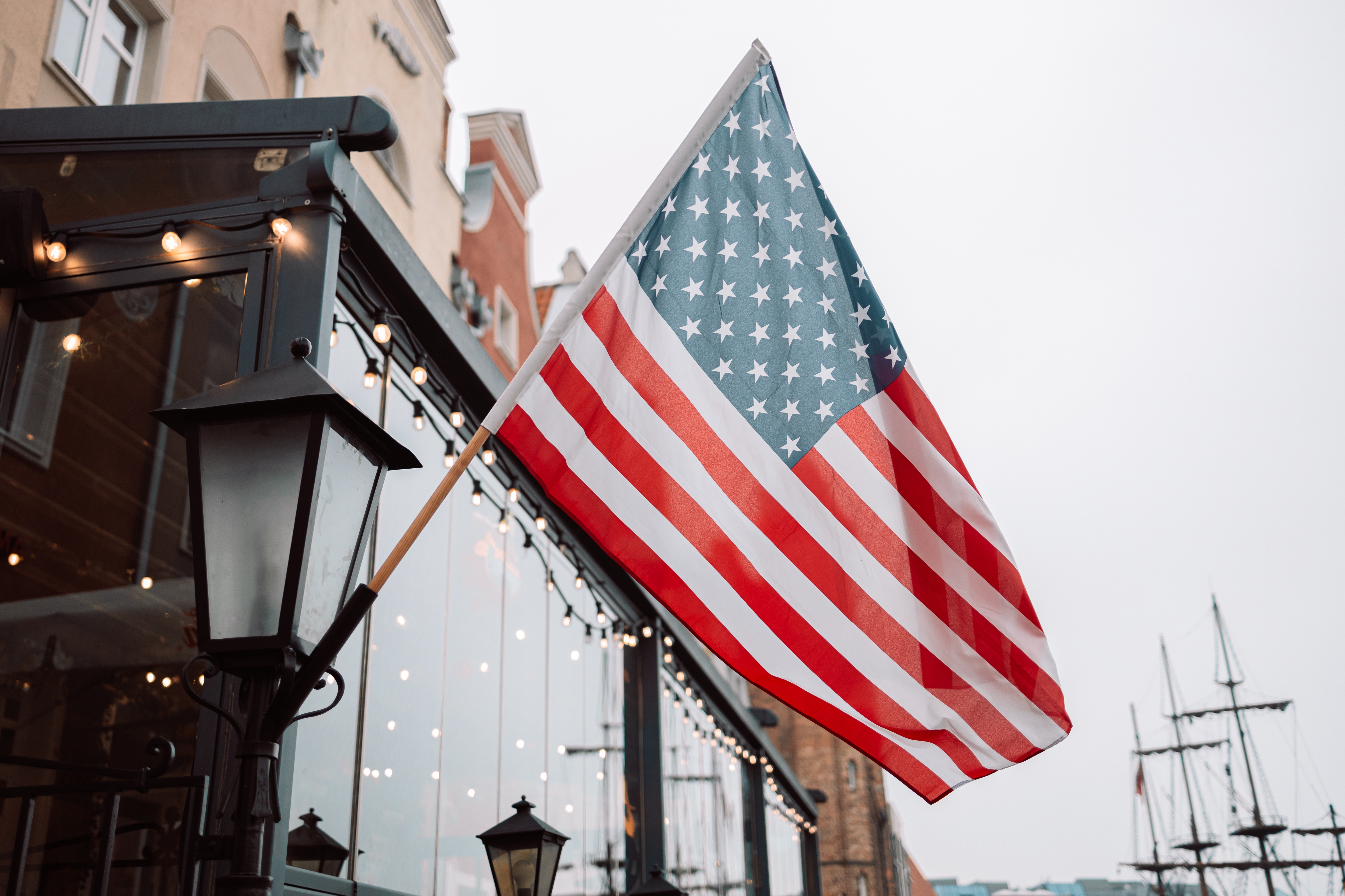 A U.S. flag waves outside a building, with ships and masts visible in the background under a cloudy sky