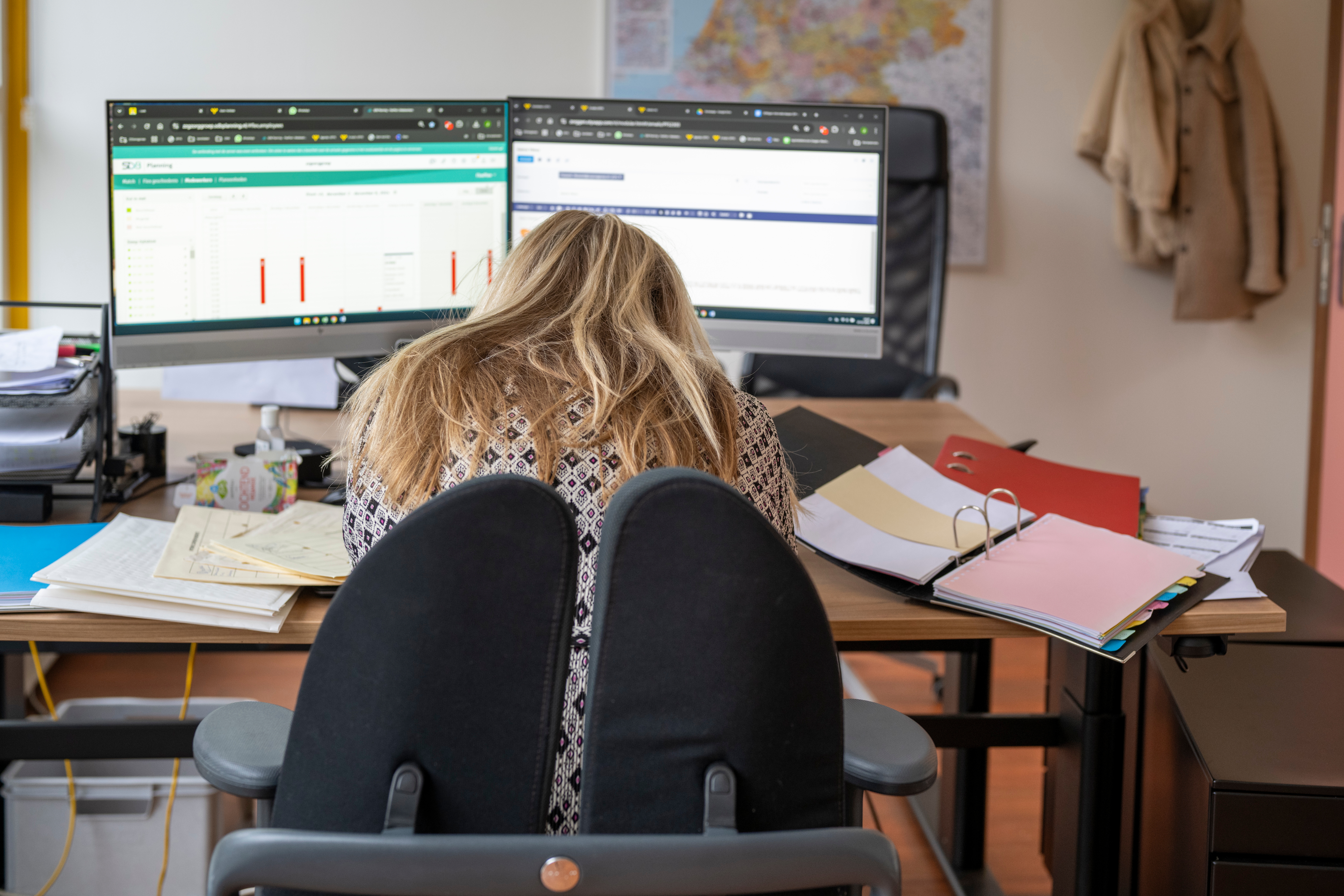 Person sitting at a cluttered desk with monitors and papers, appearing overwhelmed with work