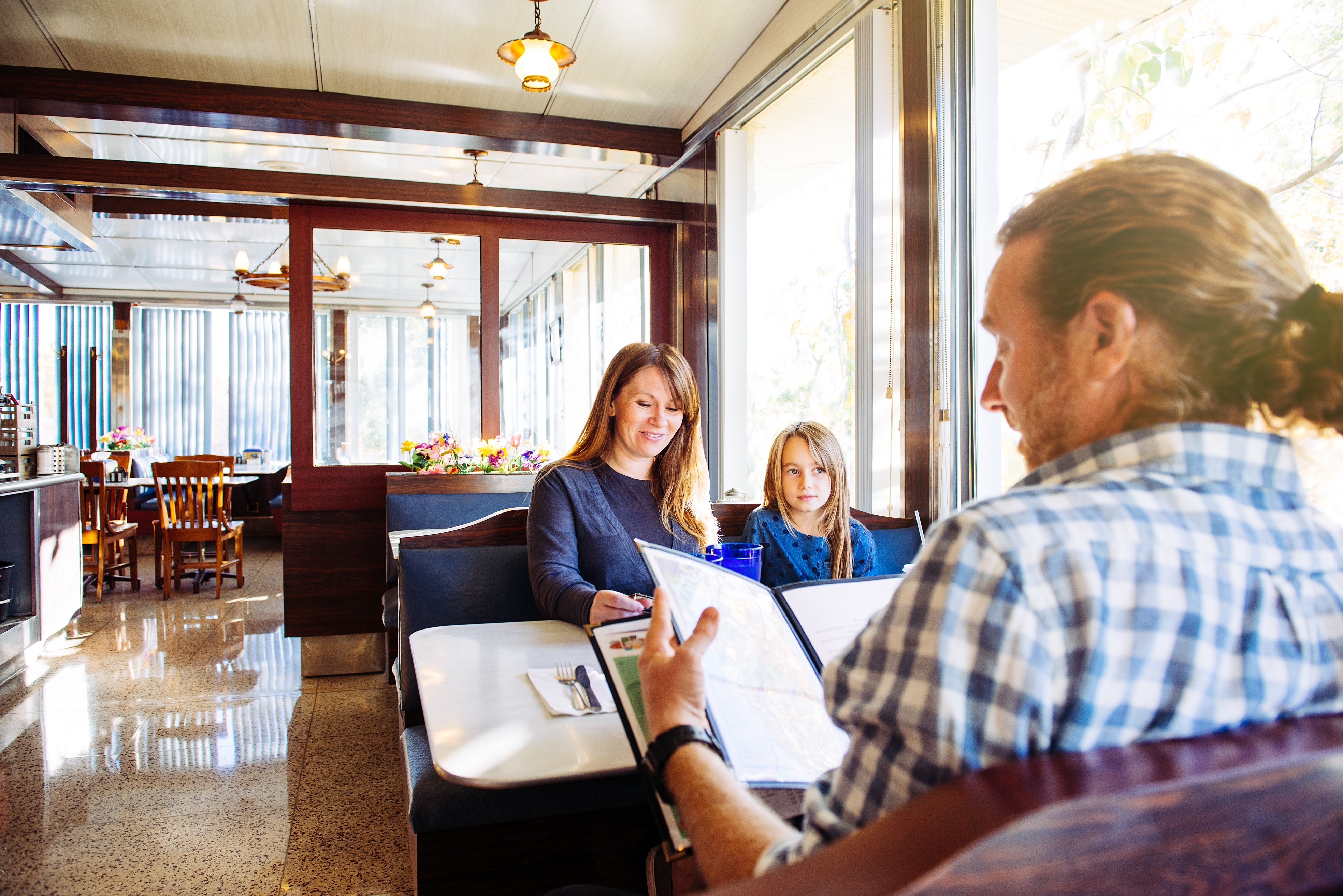 A family in a diner booth looks at menus, with a woman and child seated and a man with a ponytail across from them