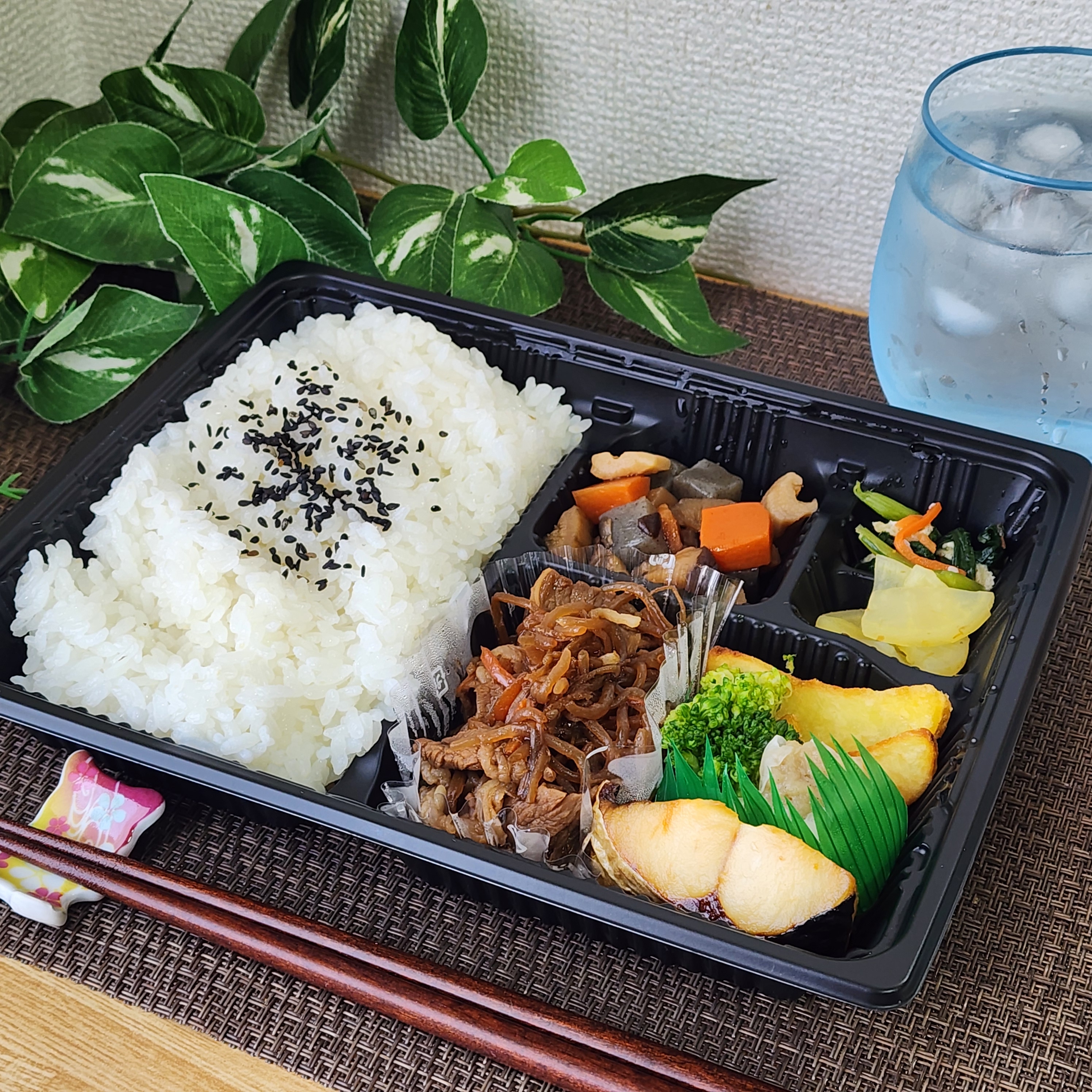 お弁当のセット:ご飯、煮物、焼き魚、漬物の盛り合わせ。冷たい水が添えられ、緑の観葉植物が背景にあります。