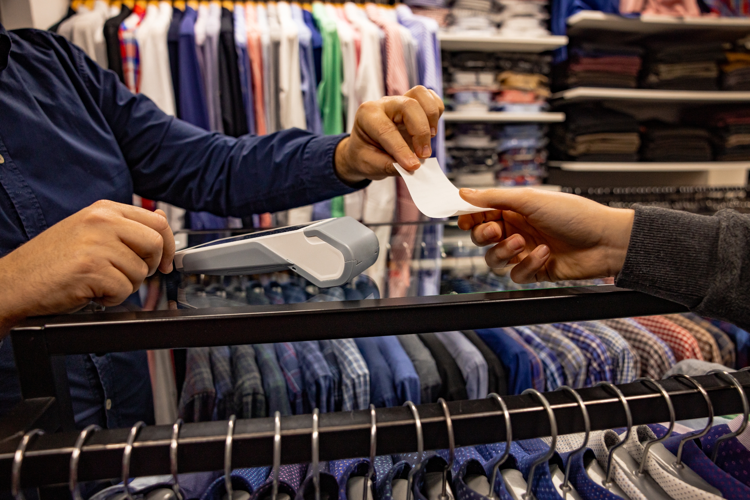 Person hands receipt to another at a clothing store, with shirts displayed in the background