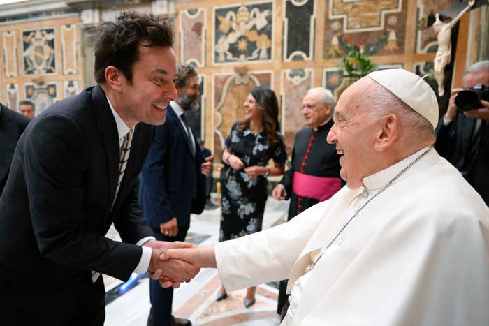 A celebrity in a suit warmly shakes hands with a religious leader in white robes and a skullcap at an ornate indoor event with other attendees