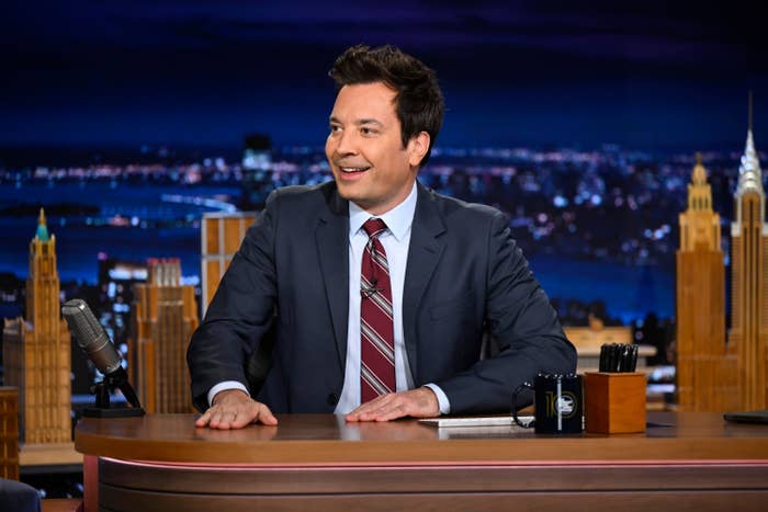 A person in a suit and tie sits at a desk on a talk show set, smiling and looking to the side. The background shows a cityscape