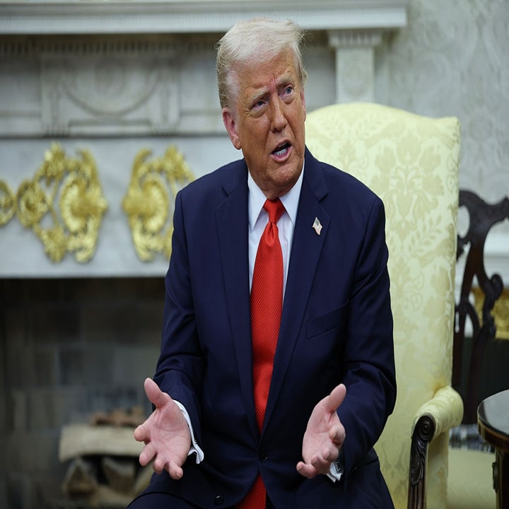 A person in a suit sits in a formal room with ornate decor, gesturing with open hands during a conversation