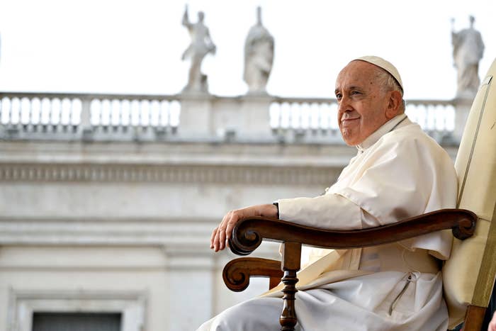 A dignified older man in religious attire sits gracefully on a wooden chair, with statues visible in the background