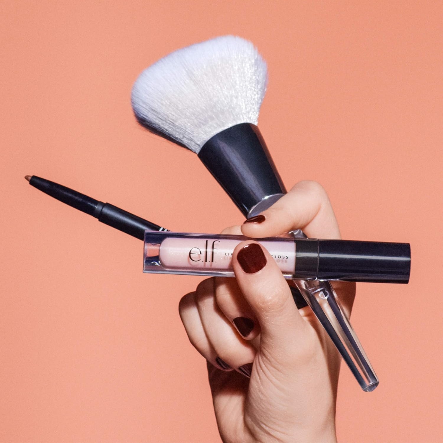 Hand holding makeup brush, eyeliner, and e.l.f. lip gloss against a plain background. Perfect for a makeup shopping article