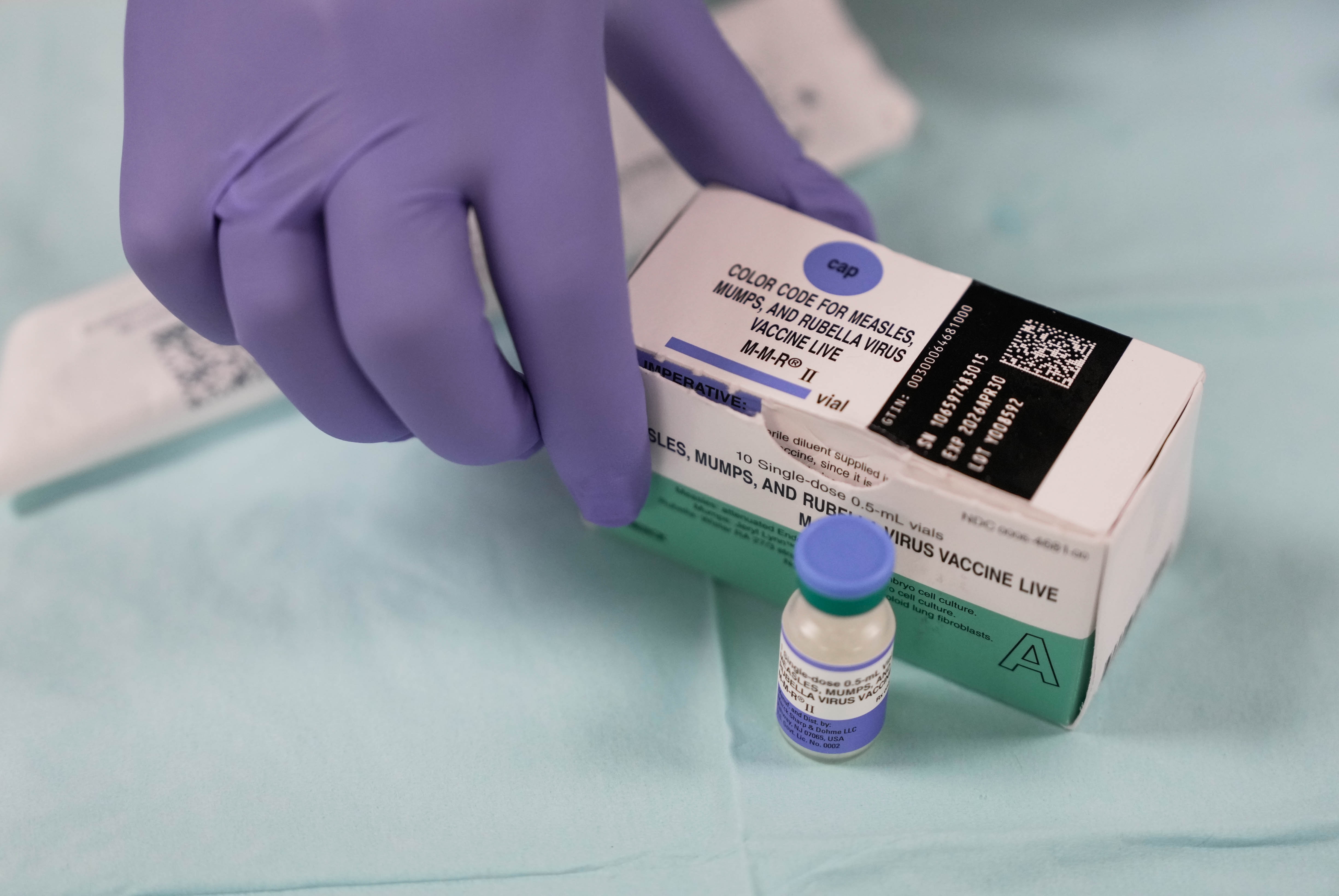 A gloved hand holding a box and vial of the MMR vaccine on a medical table