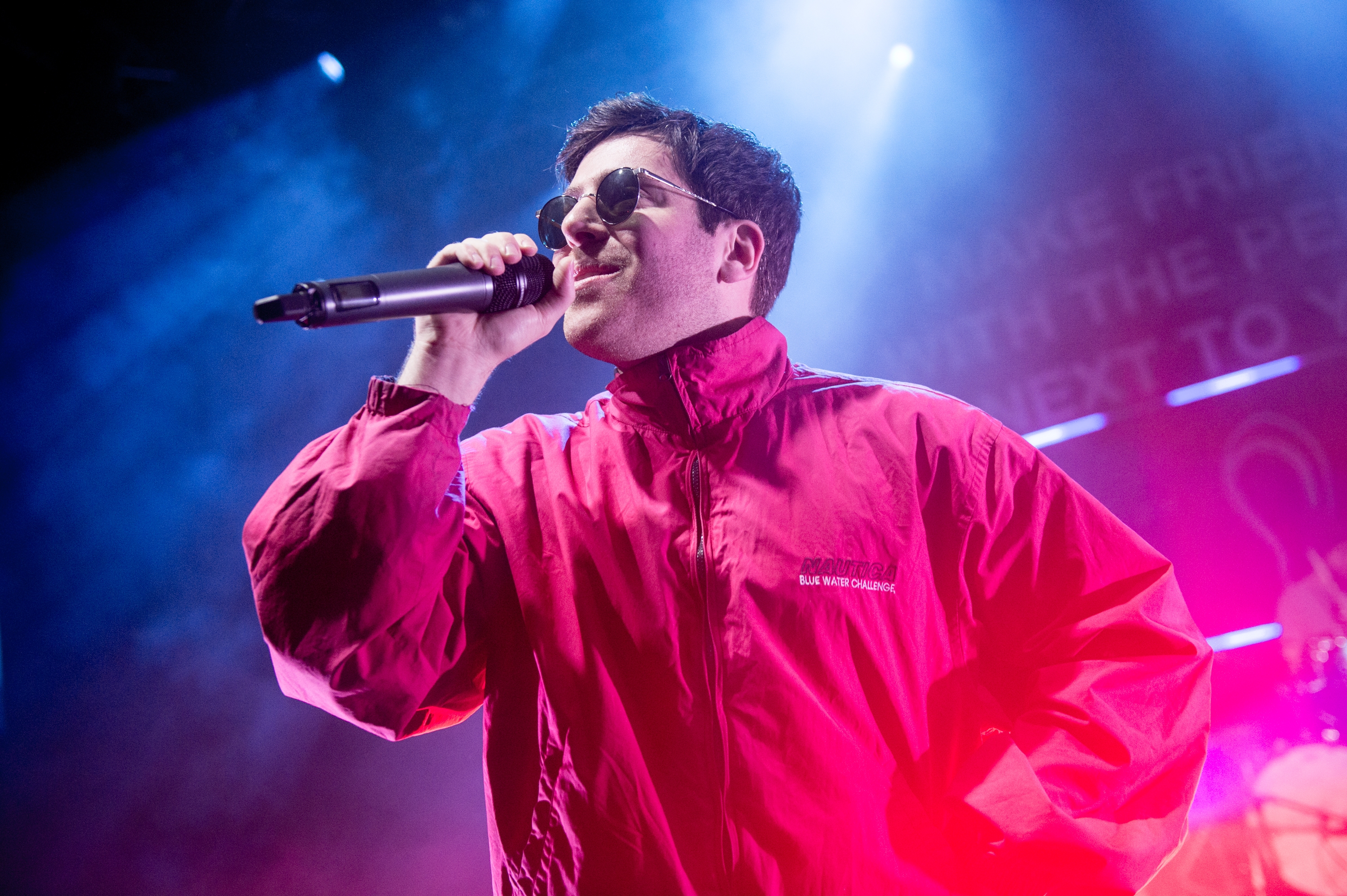 Person in a jacket and sunglasses sings into a microphone on stage with vibrant lighting