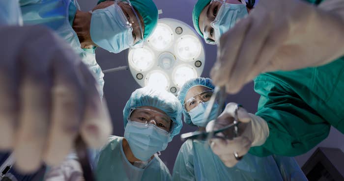 Surgeons in masks and gowns perform an operation, viewed from the patient's perspective, under bright operating room lights