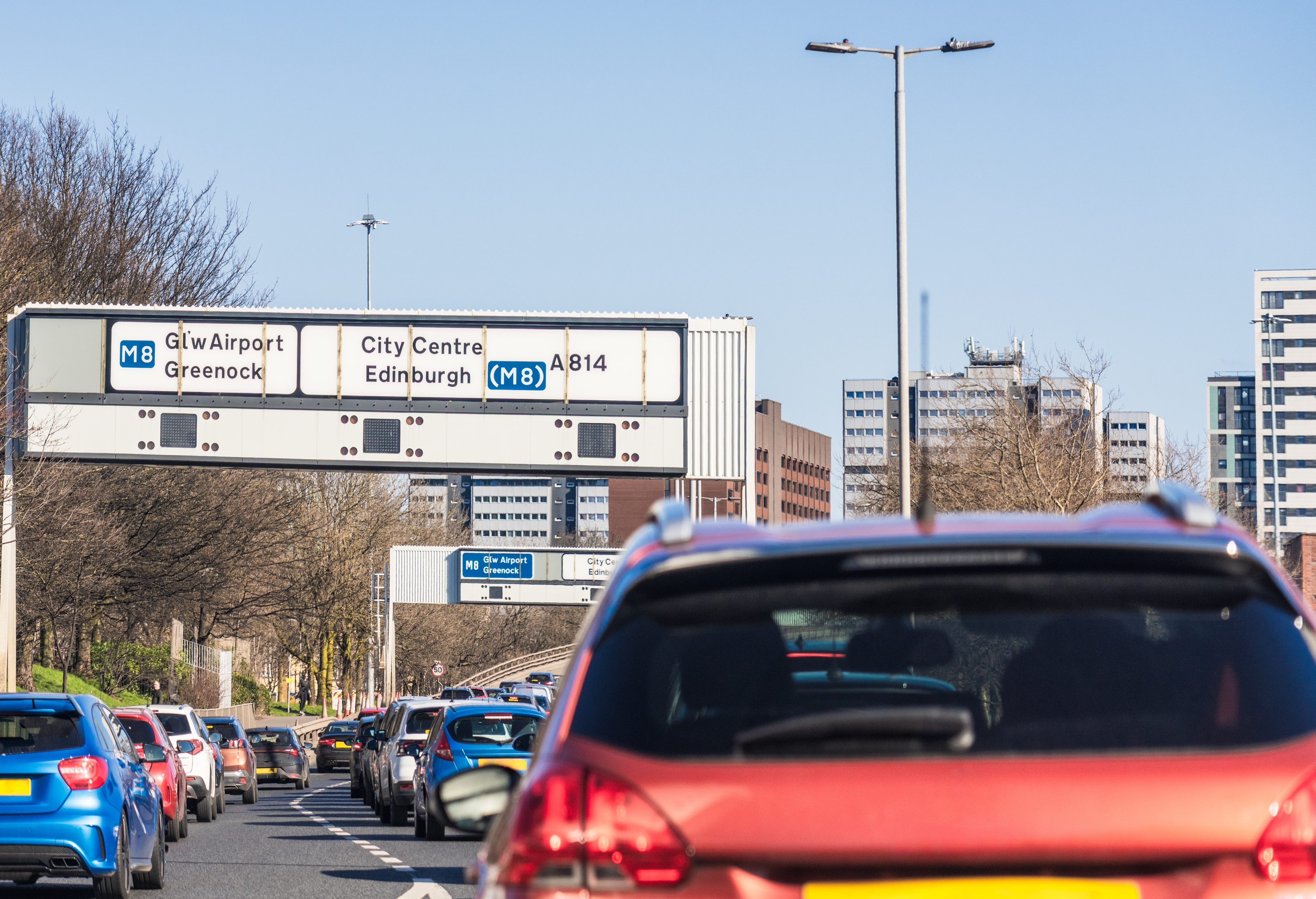 Traffic jam on a busy city road with direction signs overhead showing routes to the airport, city center, and Edinburgh