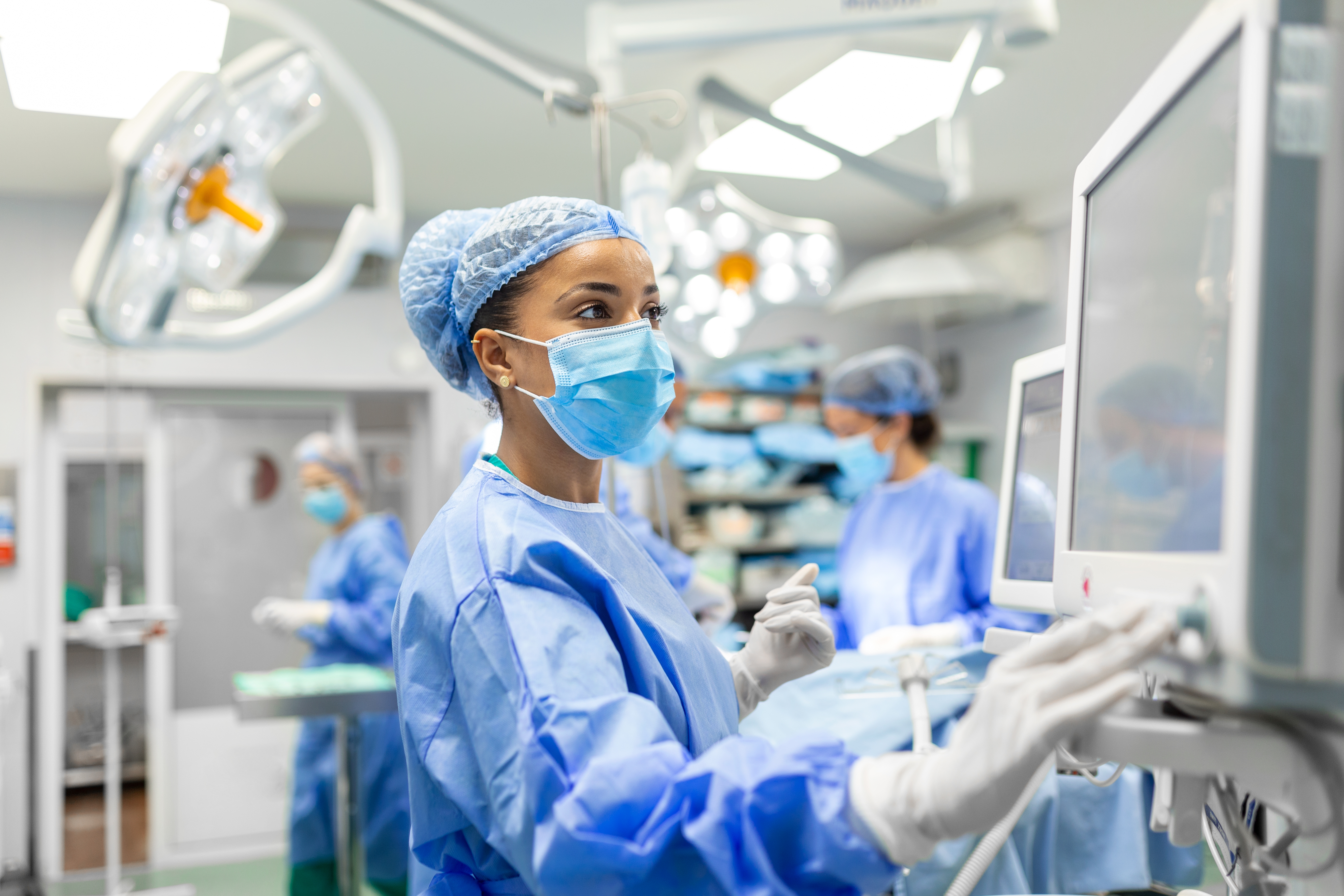 A medical professional in surgical attire operates monitoring equipment in an operating room, focused on patient care