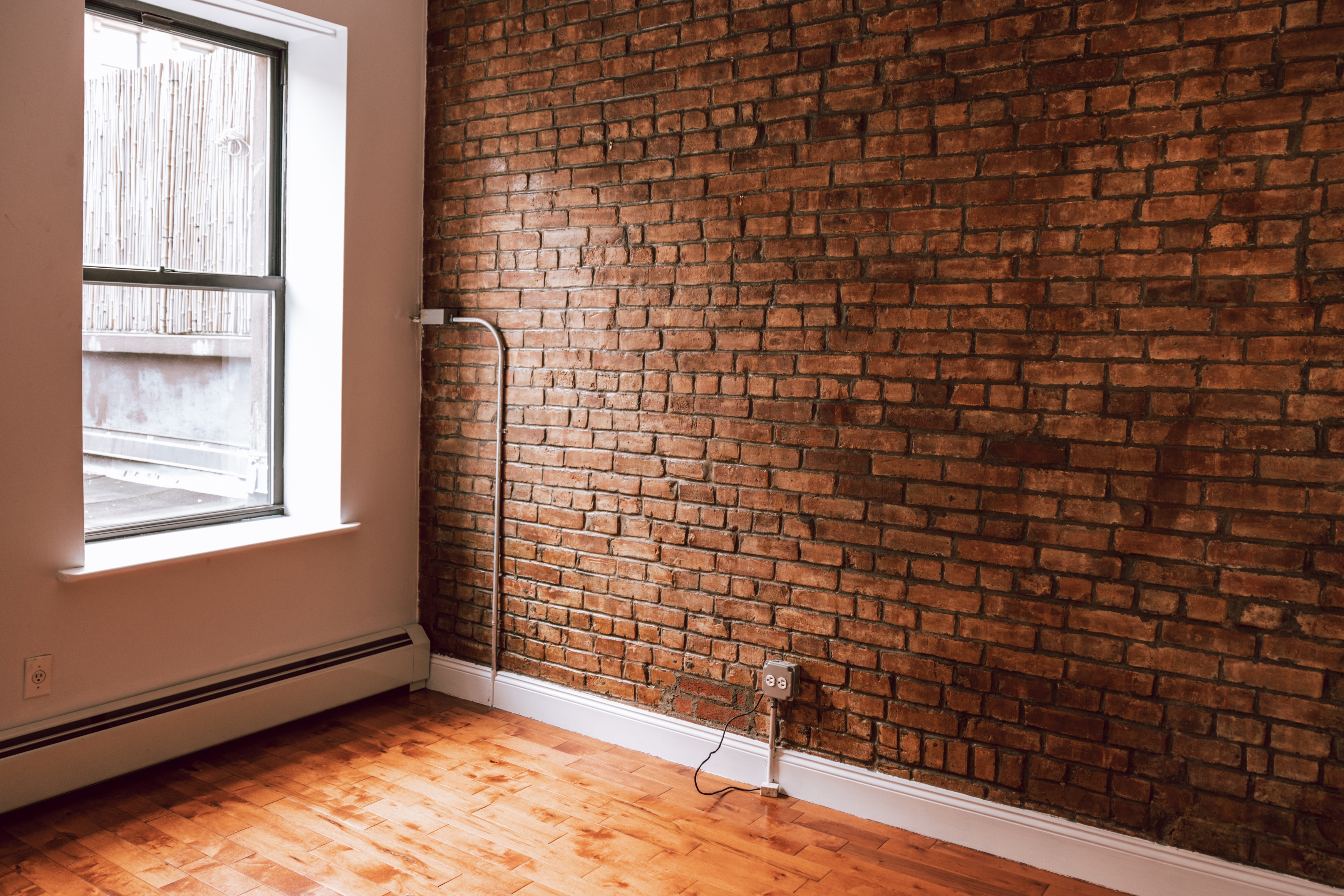 Empty room with a brick wall, wooden floor, and a window with natural light. Minimalist and rustic setting