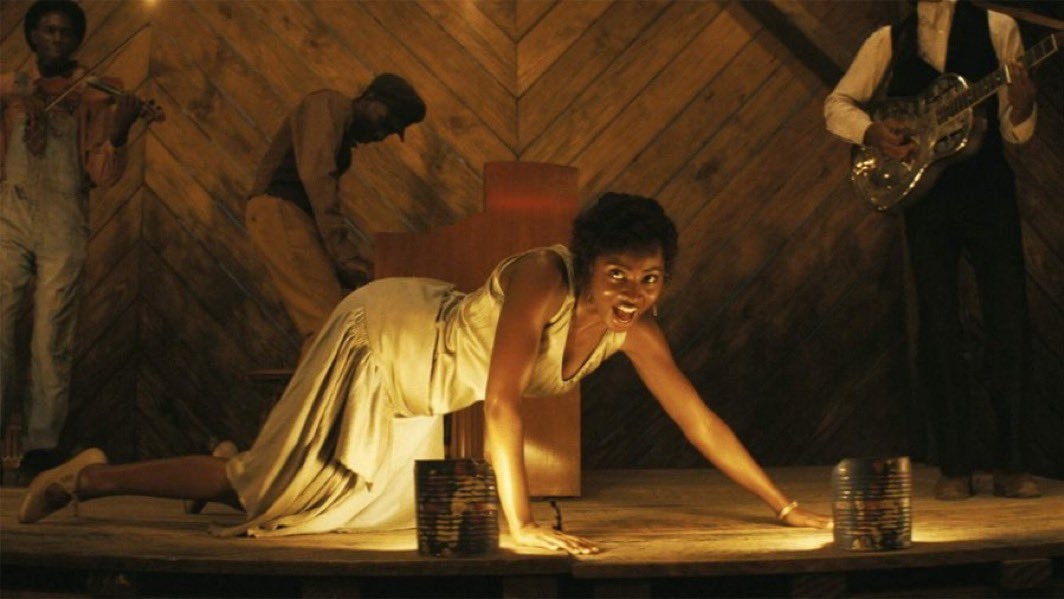 Mujer en pose teatral en un escenario tipo cabaret, rodeada de músicos. Lleva un vestido elegante y expresivo