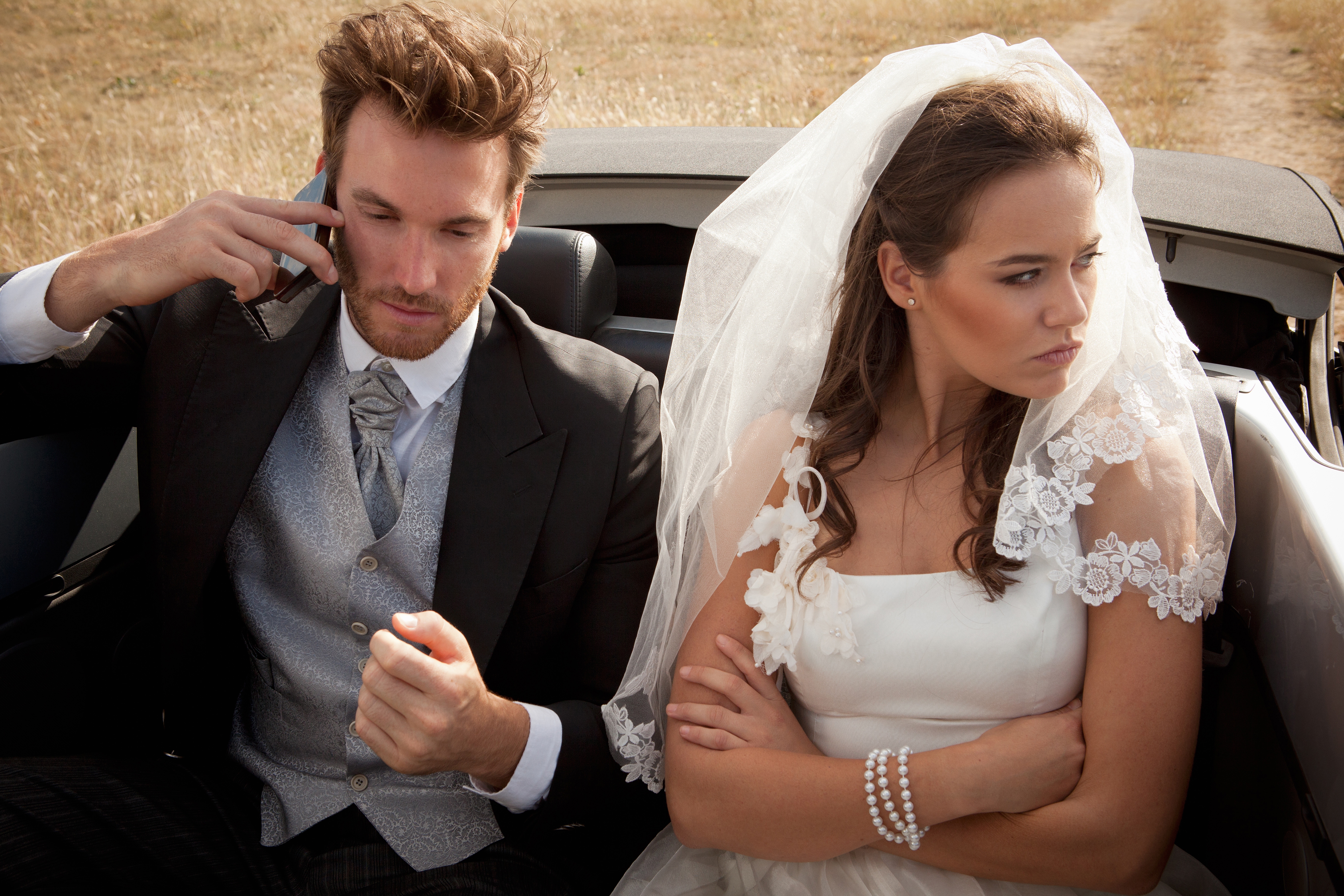 Bride in a white lace dress looks upset, sitting next to a groom in a tuxedo talking on a phone. They appear to be in a convertible