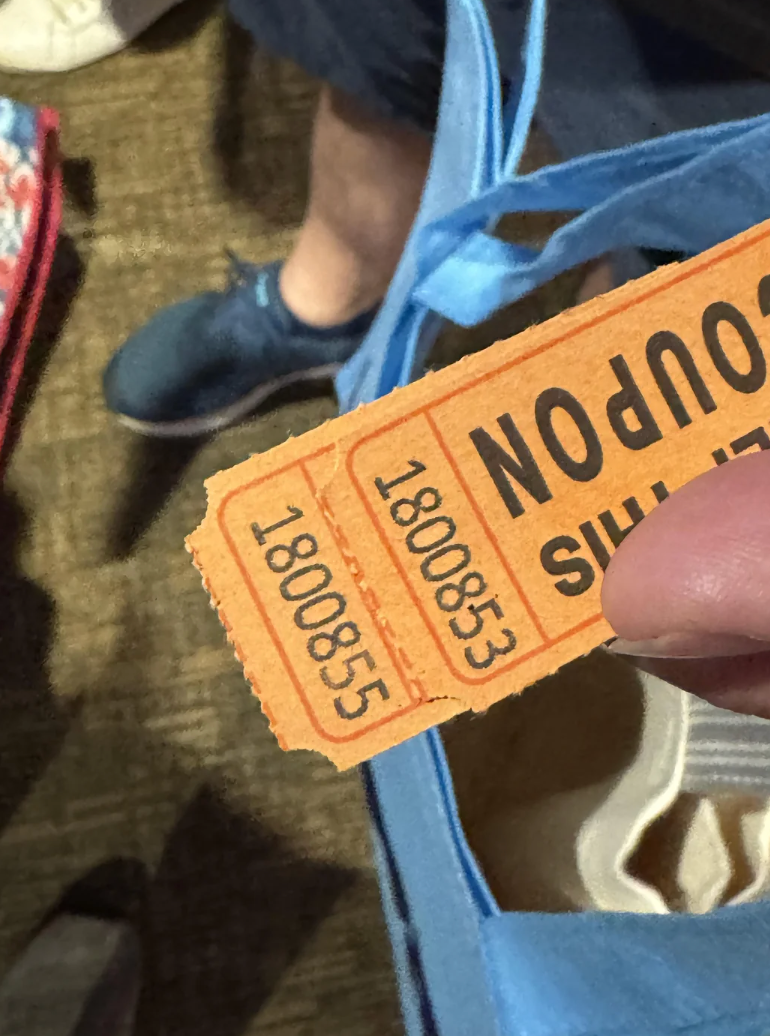 Person holding a raffle ticket with the number 1800853, above a blue bag. The ticket is partially torn, indicating it&#x27;s been used