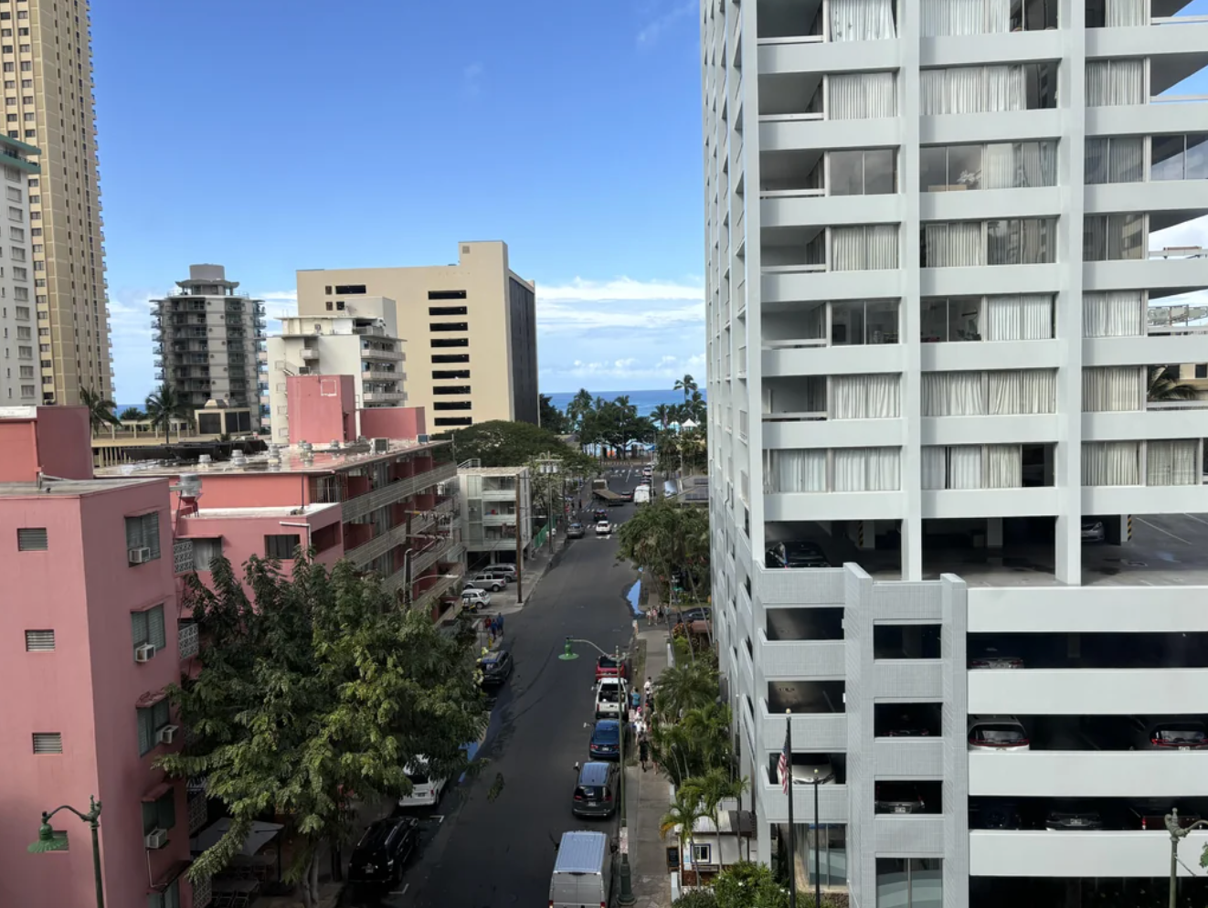 Urban street view with tall buildings, trees, and cars. A glimpse of the ocean is visible in the distance