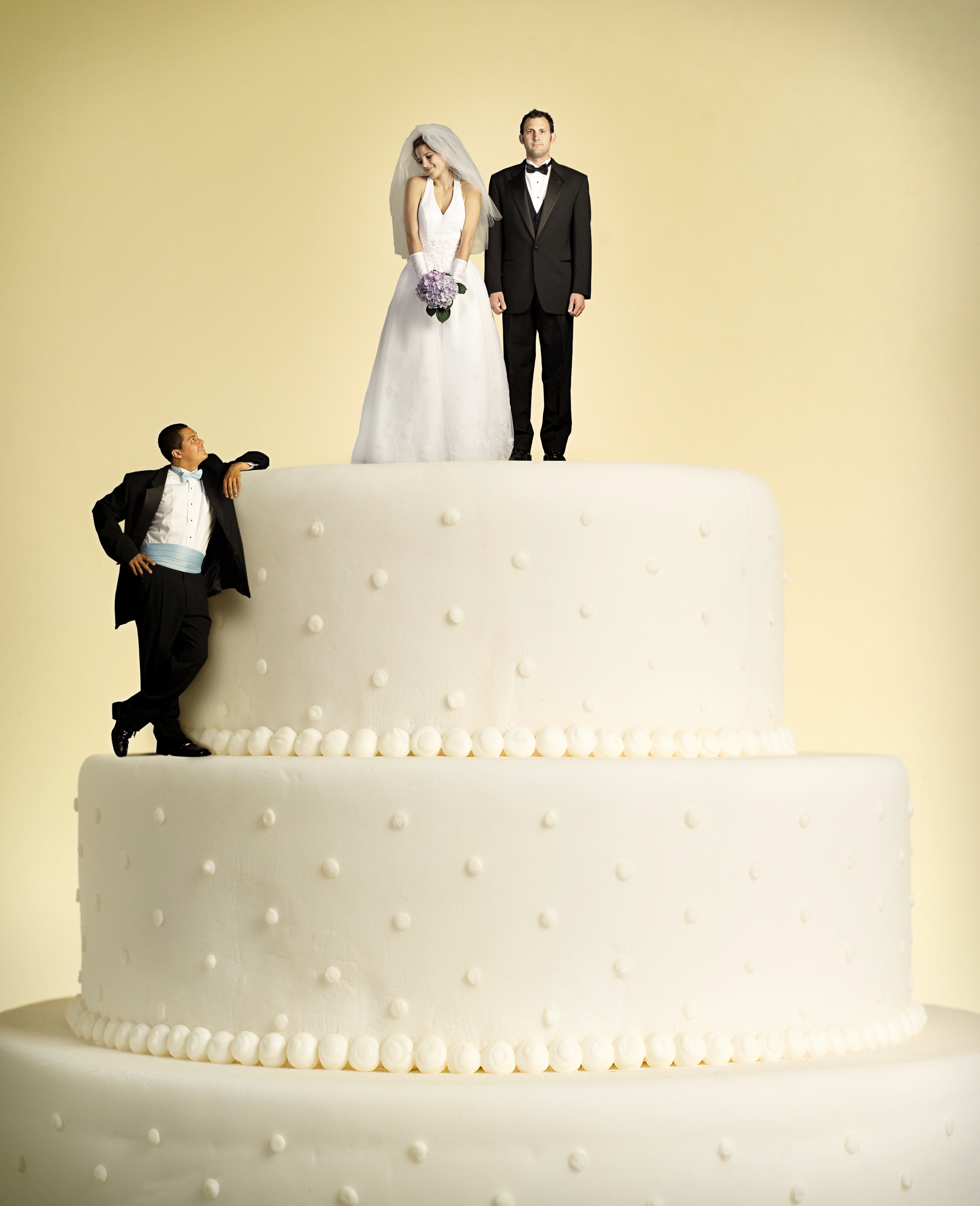 Bride and groom figurines stand atop a three-tiered cake, with a groom on the lower tier looking up at them, in a humorous wedding scene