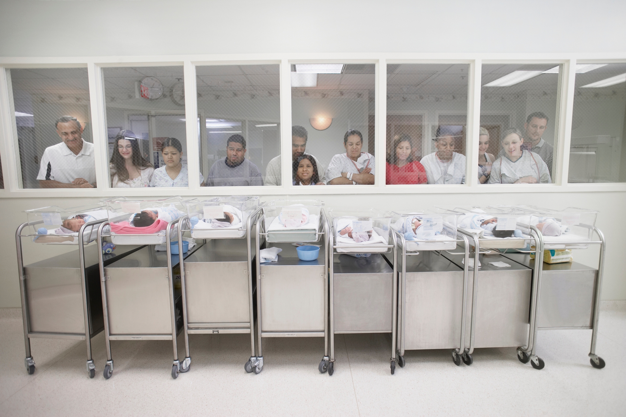 Parents look through a window at newborns in a hospital nursery, observing their babies in cribs lined up in a row