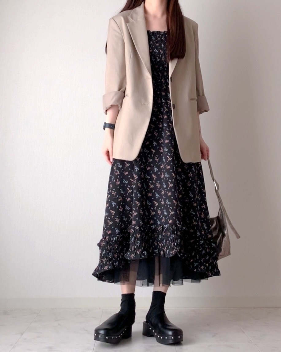 白い背景の前で、花柄ドレスにベージュのジャケットとブーツを合わせた女性が立っている。シンプルでシックなファッション。