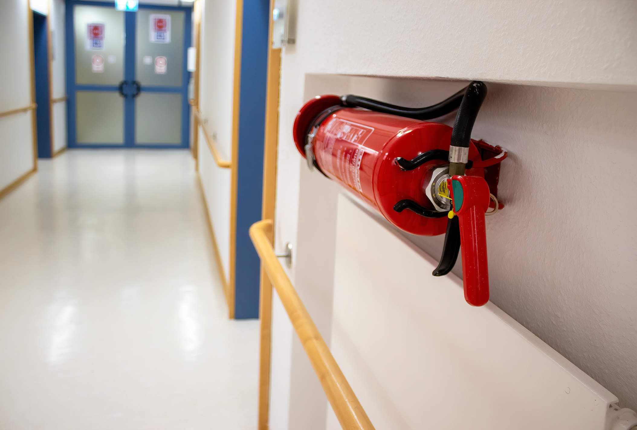 A fire extinguisher mounted on a wall in a school hallway, with doors and safety signs visible in the background