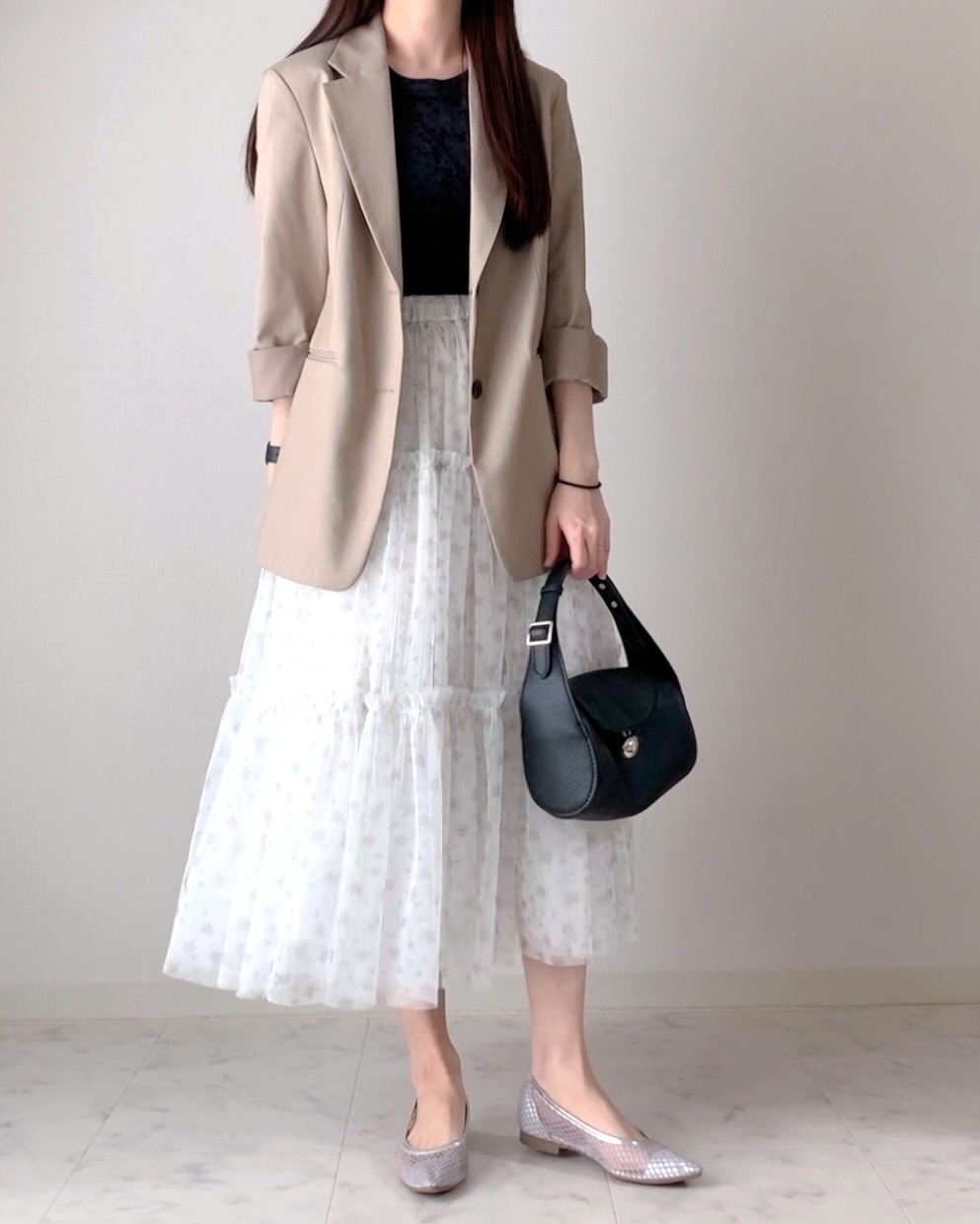女性がベージュのブレザーと白いロングスカートを着用し、黒いバッグを持っています。