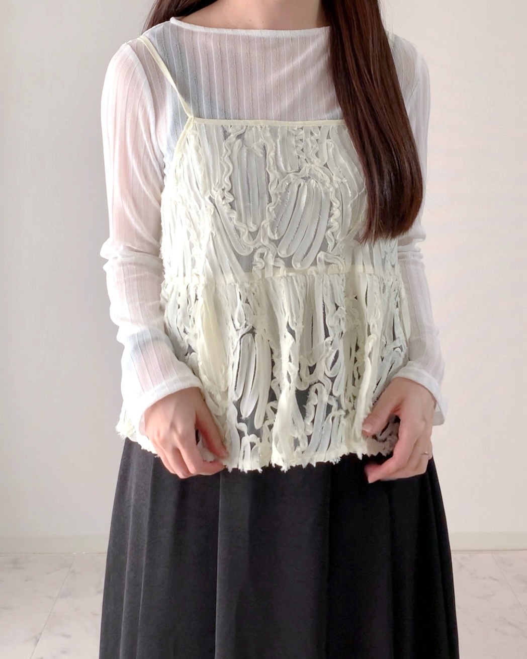 Person wearing a sheer, long-sleeve top layered with a textured, fringed camisole and a dark skirt. Face not visible