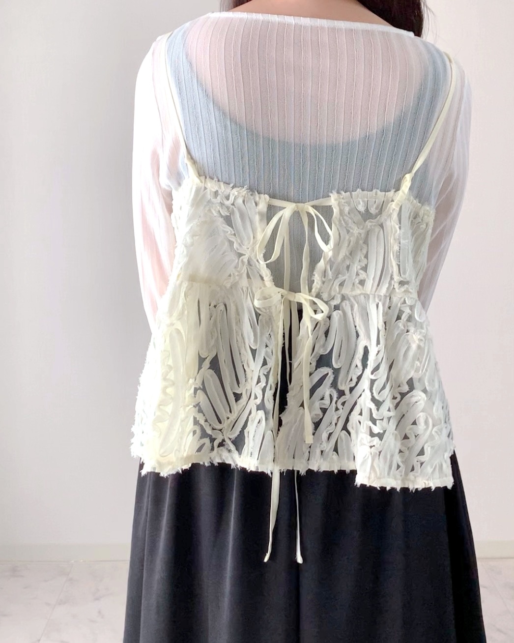 A person is shown from the back wearing a layered outfit with a sheer white lace top over a long-sleeved shirt and a dark skirt