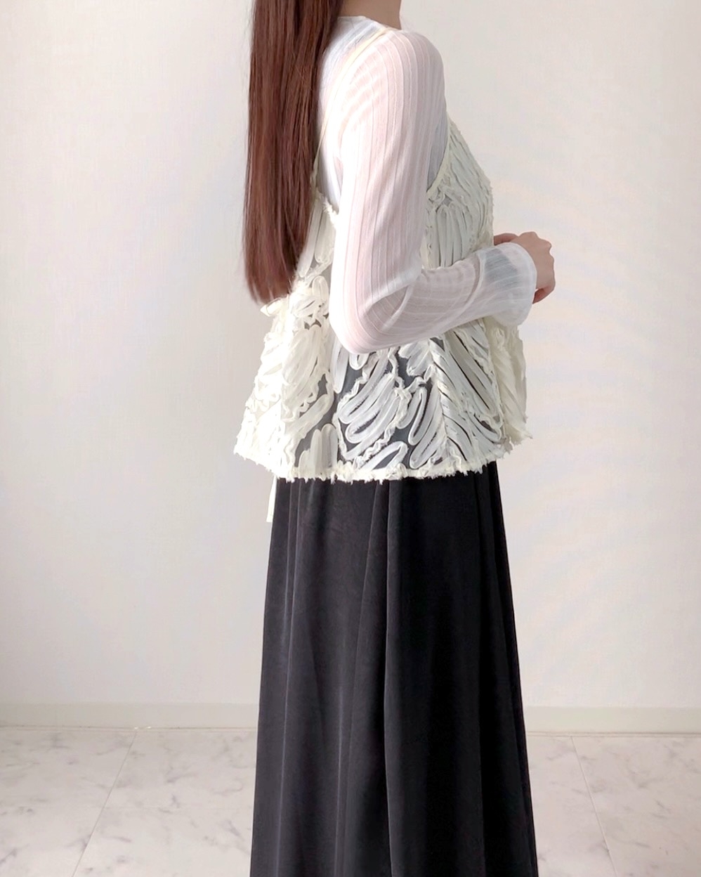 Person with long hair wearing a layered outfit with a textured top and flowing skirt, standing against a plain background