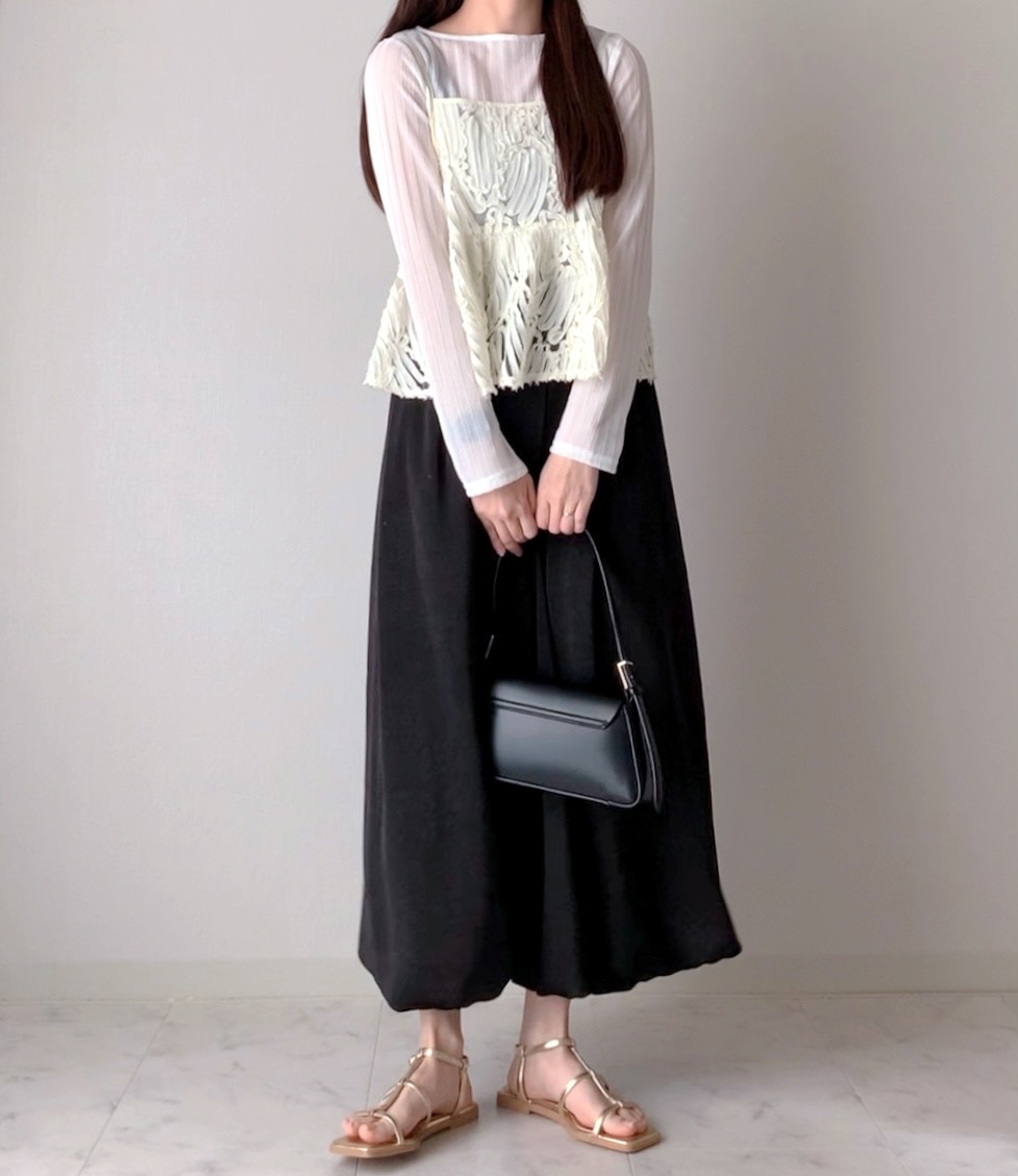 A person wearing a sheer white top with a lace overlay, black skirt, strappy sandals, and holding a black handbag, stands against a plain backdrop