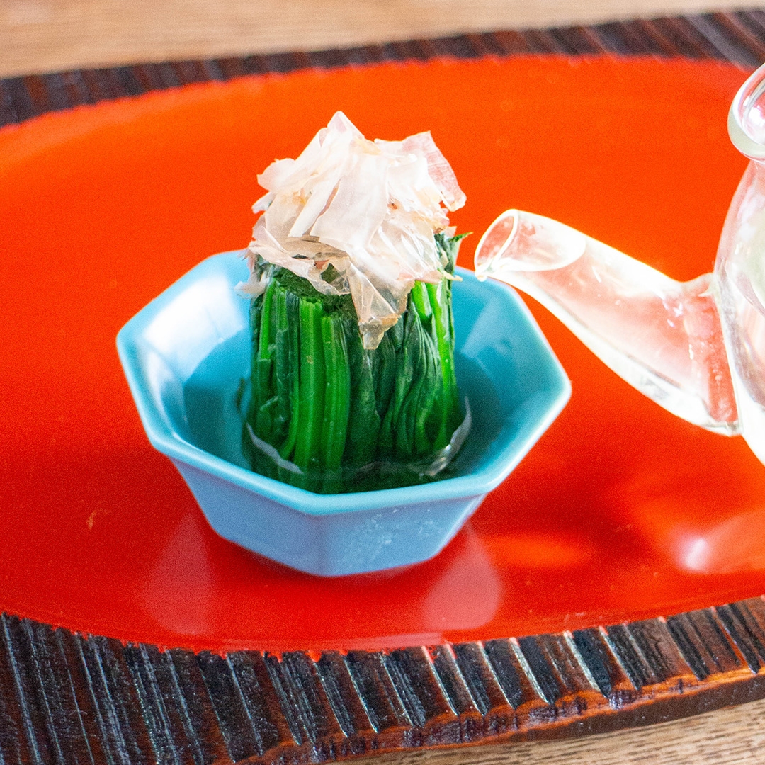 青い器に盛られたほうれん草のおひたし、上にかつお節がのっている。横に透明なポットが置かれている。