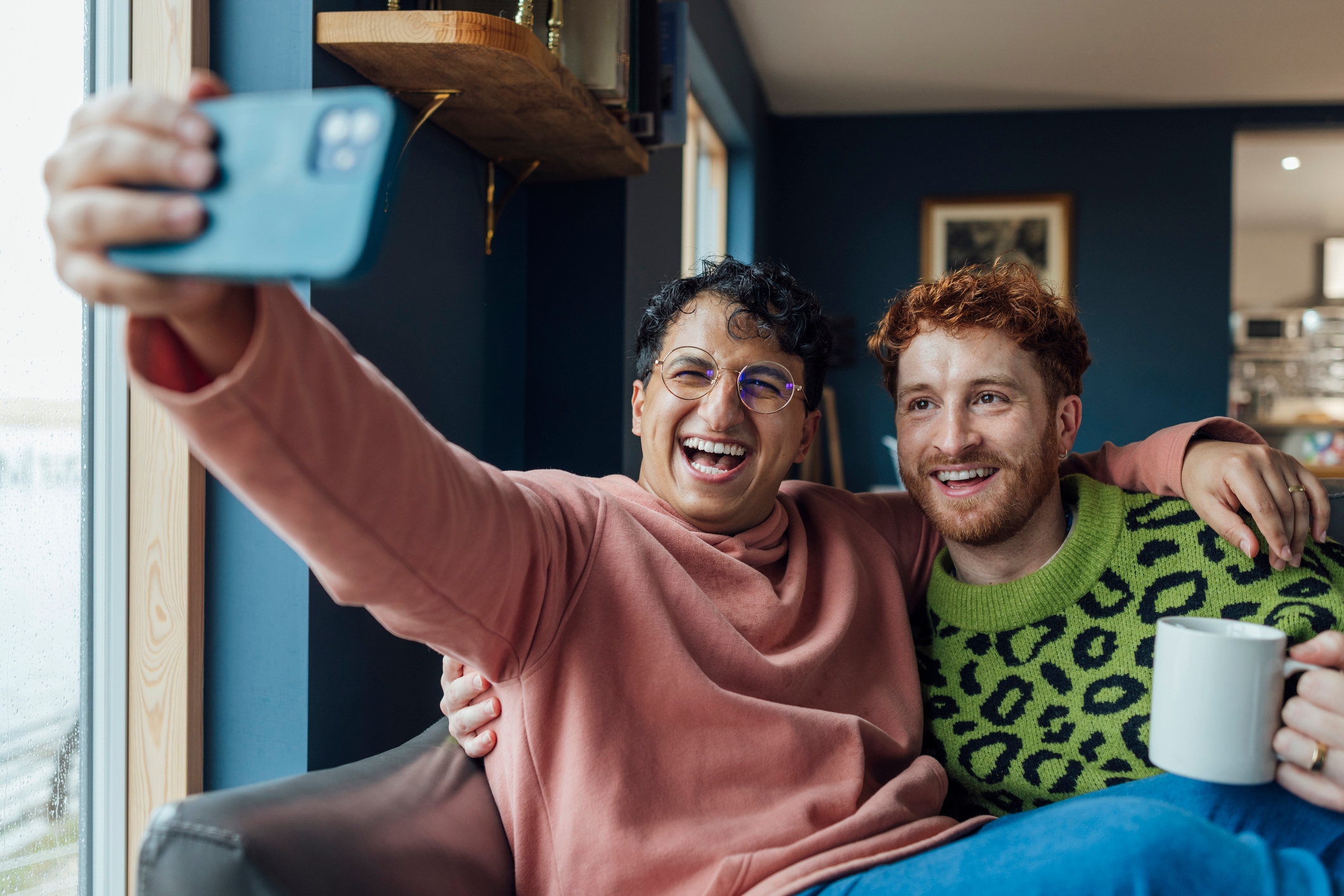 Two friends smiling and taking a selfie; one wears a sweater, and the other, holding a mug, wears a patterned sweater